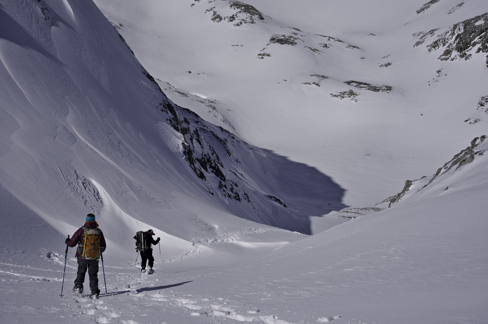 Two people descend a steep snow field. A steep mountain side is on the left partly in shadow