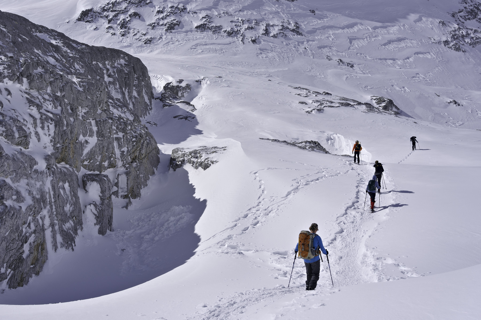 A line of people in snowshoes pass to the right of a large cliff face covered in snow and ice