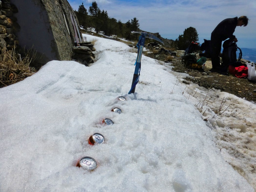 A row of beer cans lies cooling in the snow. An ice axe stands beyond them and a person sorting out a rucksack to the right.