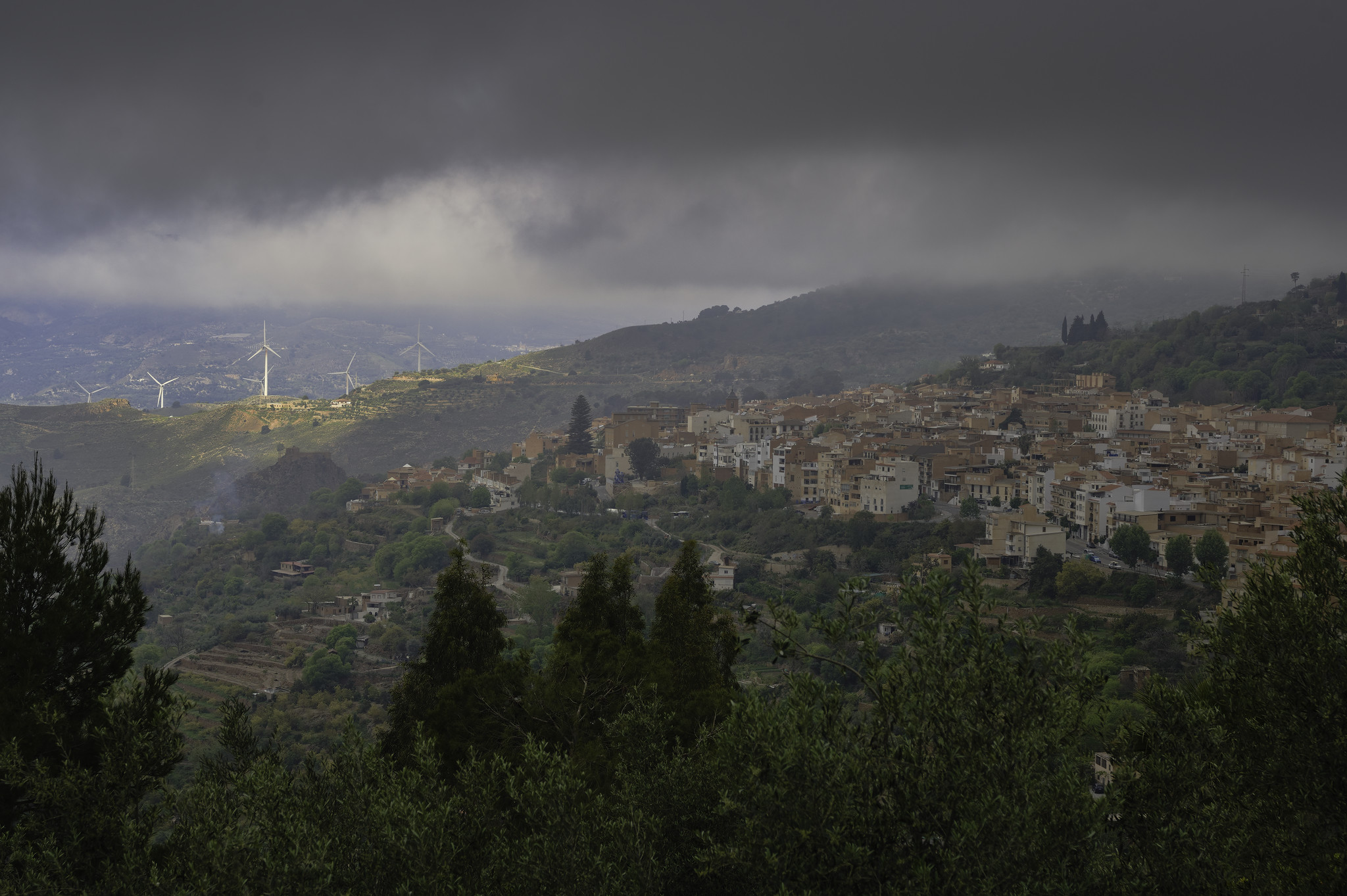 Some gray clouds hang over a small town set on a hillside to the right. To its left there are some wind turbines illuminated by a weak sun. In the foreground some olive trees