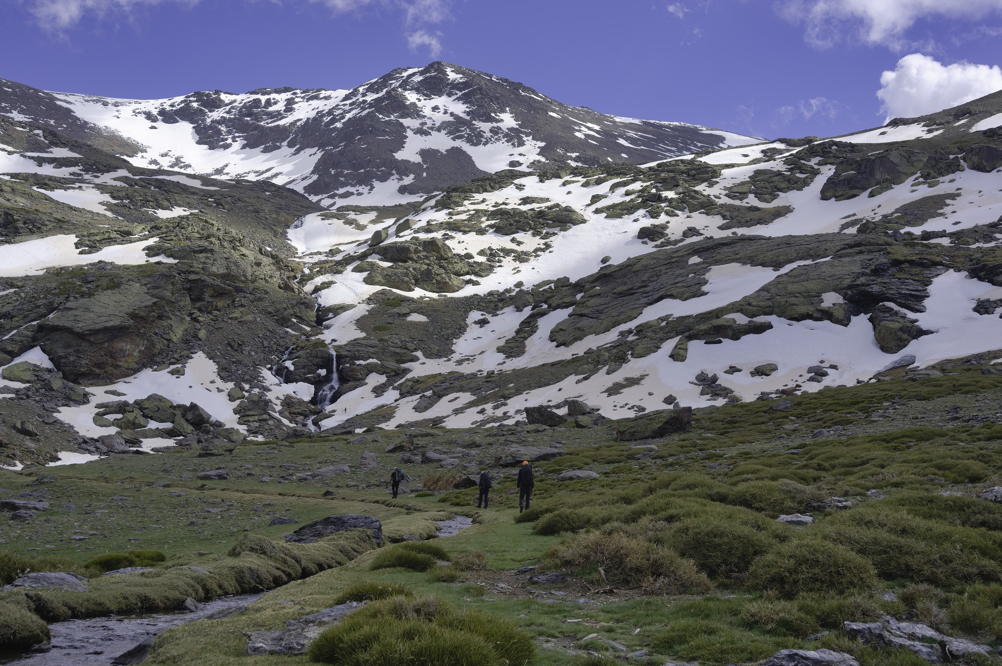 3 backpackers hike in along a green path towards a valley where the snows start and a waterfall can be seen. The valley rises to a high peak in the background