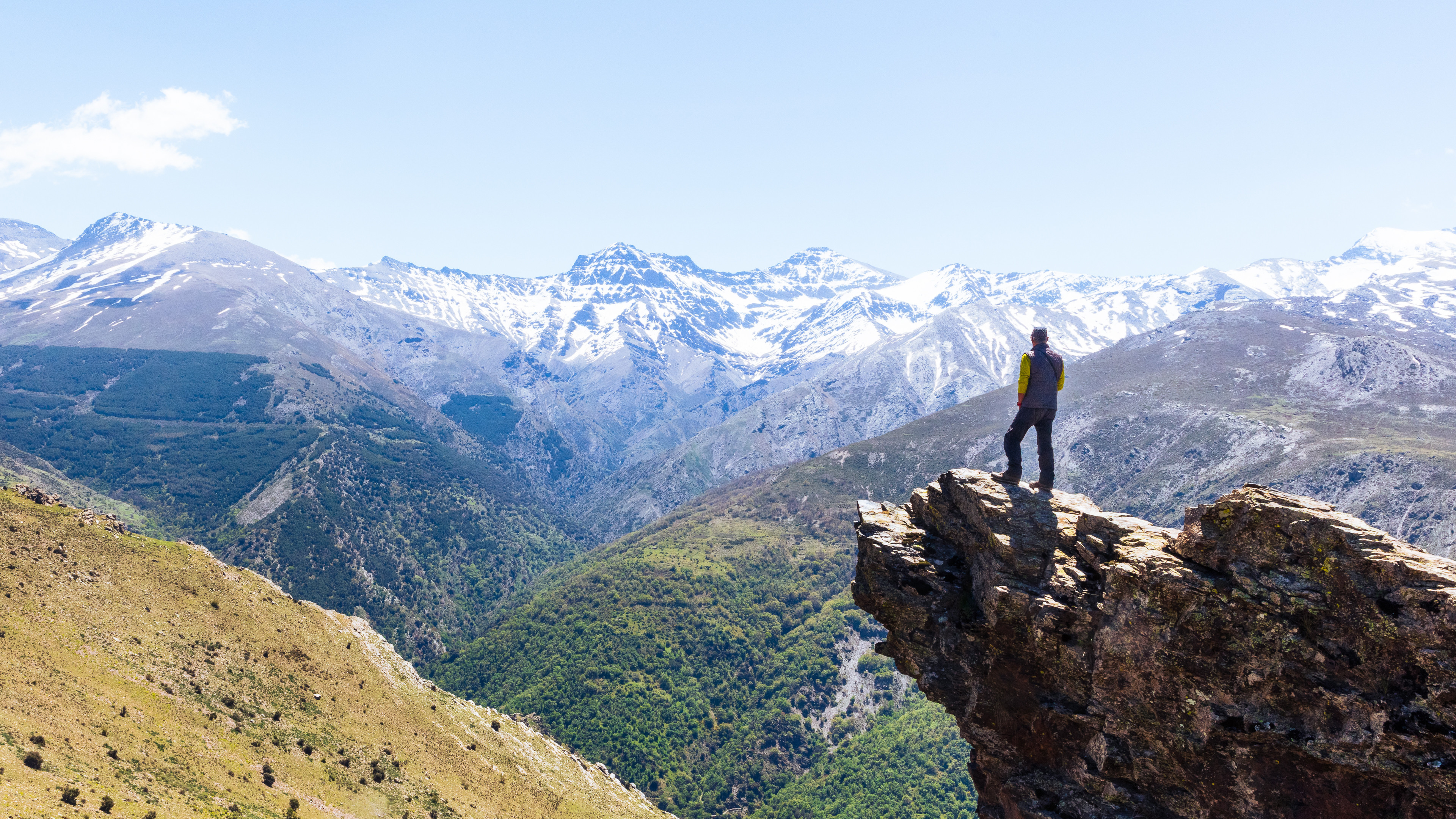 A person (me!) stands on a rocky outcrop above a void. In the background yellow and green valleys give way to snow covered mountain tops.