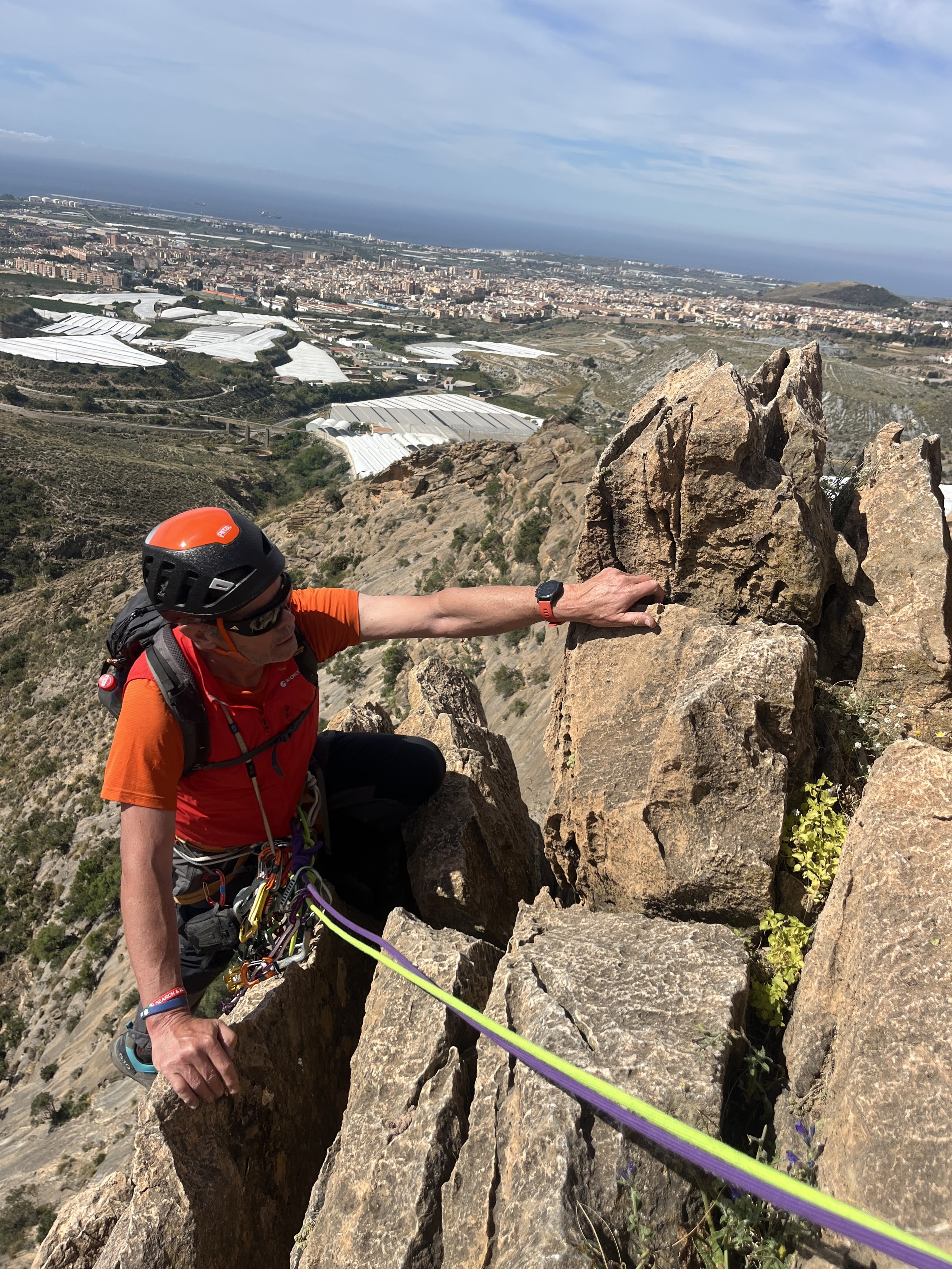A climber in red grips the orange rock. In front of him are two ropes. In the distance a city and the sea beyond