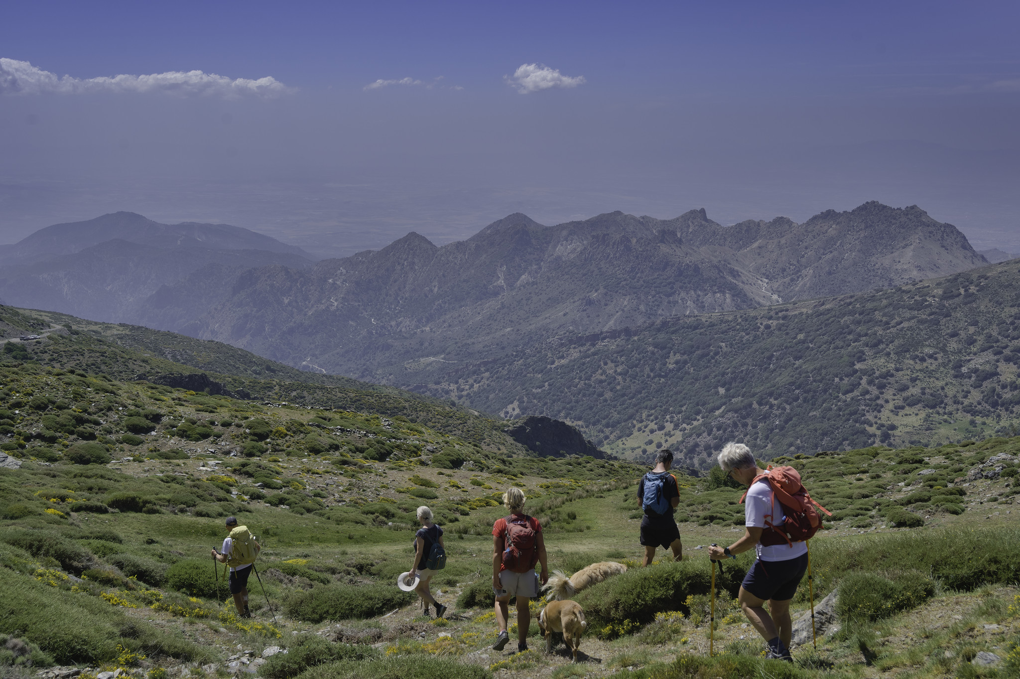 Hikers descend some green and yellow slopes. The valley ahead converges to some distant mountains. Blue sky above