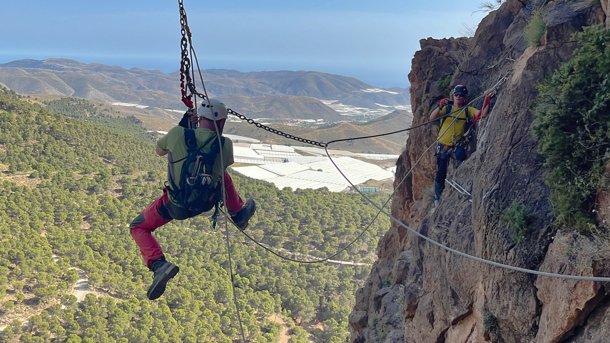 A person swings out from a mountain face on a pendulum swing. behind are green trees and the sea
