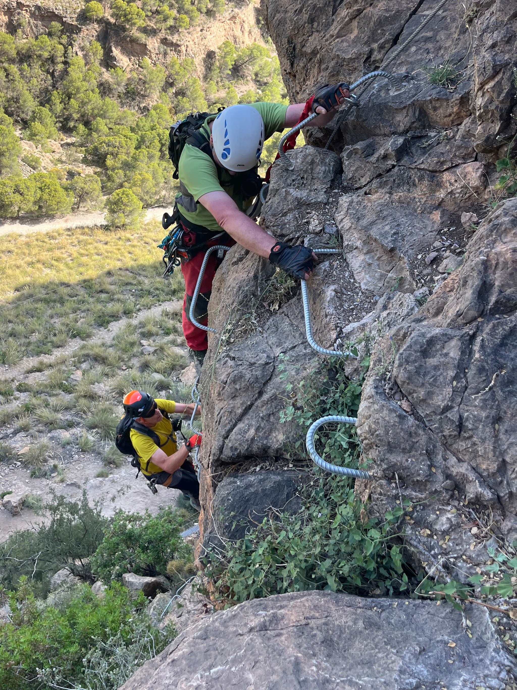 2 people climb iron rungs cemented into the rock face. Both wear helmets