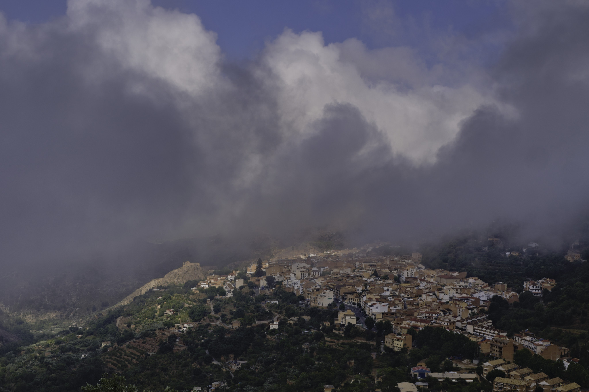 A village sits on a hill with dark clouds above and blue sky above that. Shafts of light come through the clouds to illuminate some of the village.