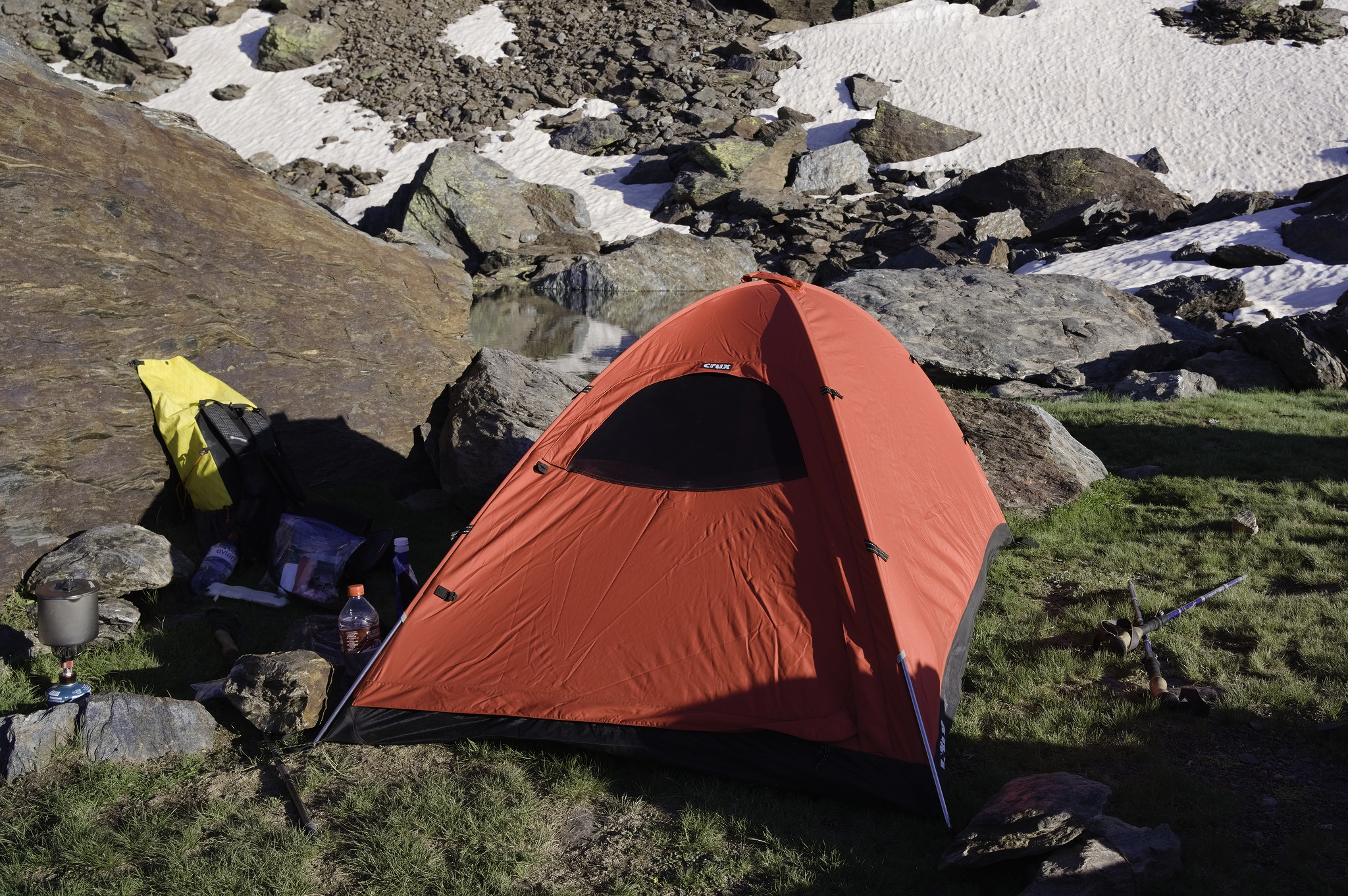 A bright red tent site next to a rock. Beyond is a small lake. near to the rock is a yellow rucksack. behind the lake are snow slopes