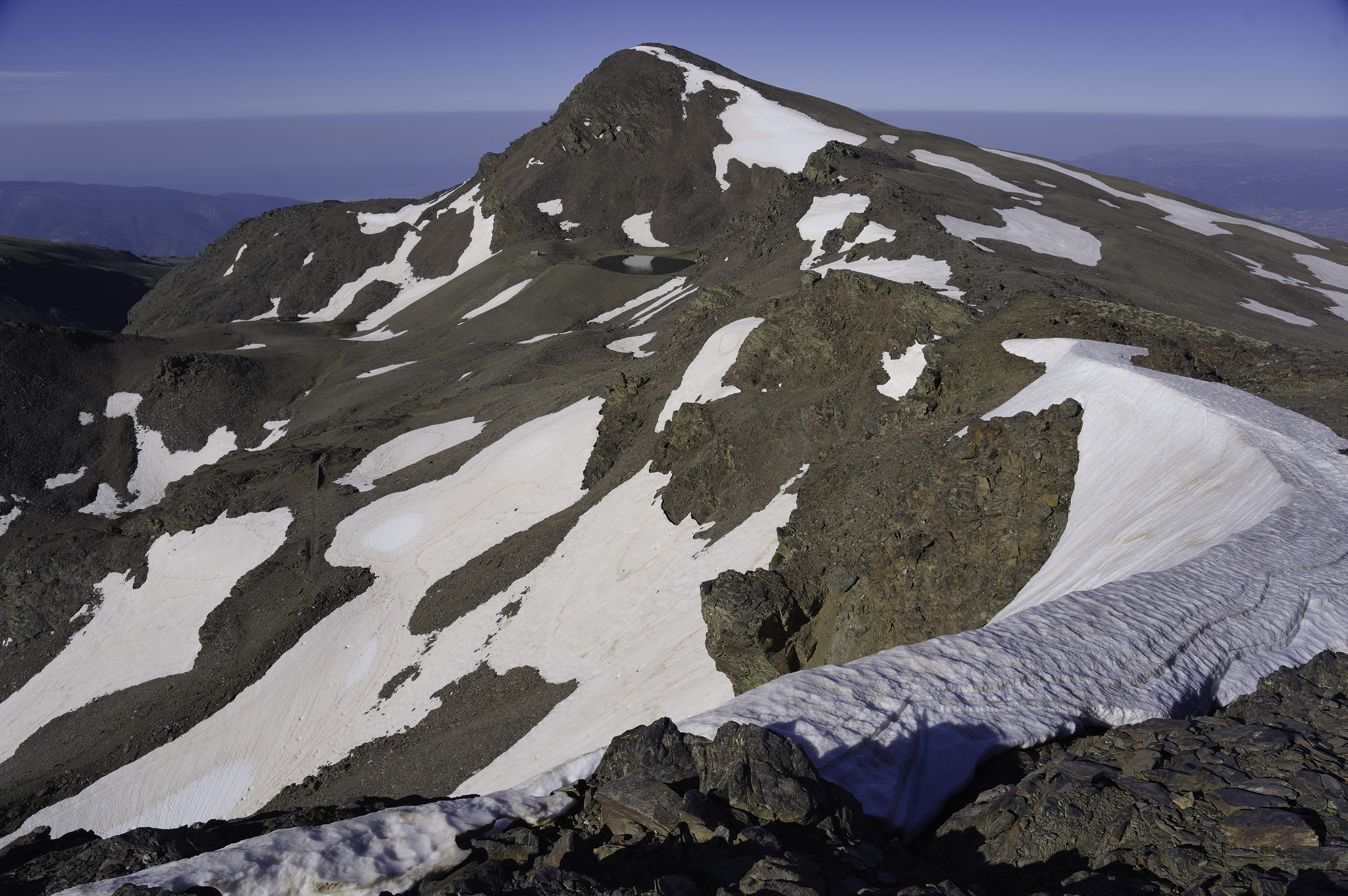 A cornice of snow curls round to the right leading to a ridge and a mountain summit. To the left snow slopes drop off. In the distance the Mediterranean sea in a heat haze