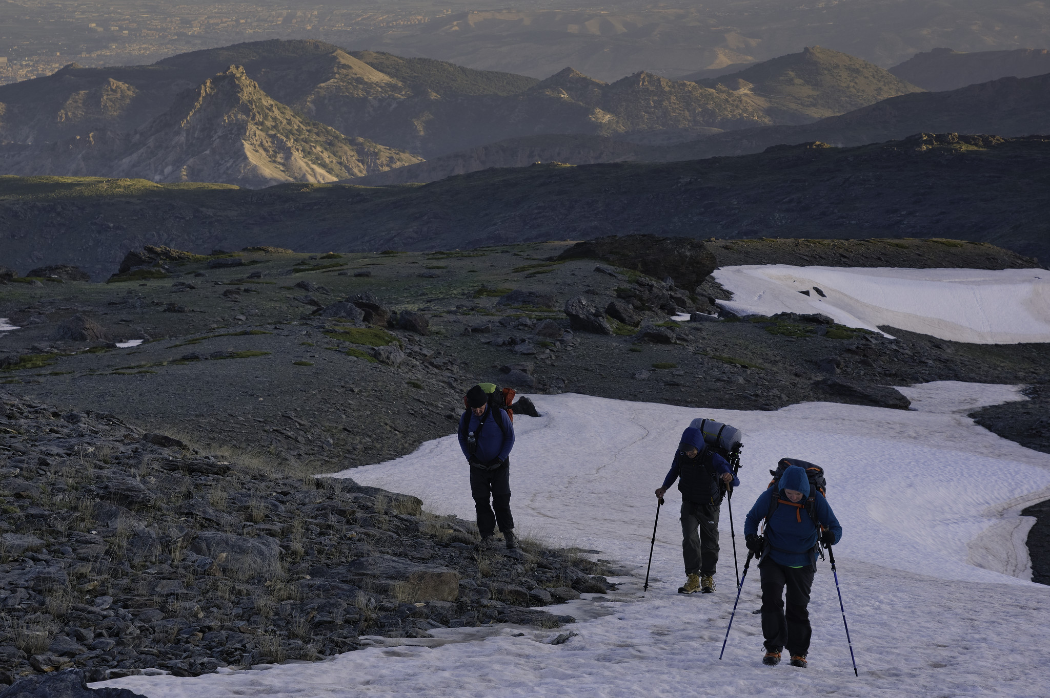 3 people, looking cold, climb a snow slope with mountains behind them illuminated by the rising sun