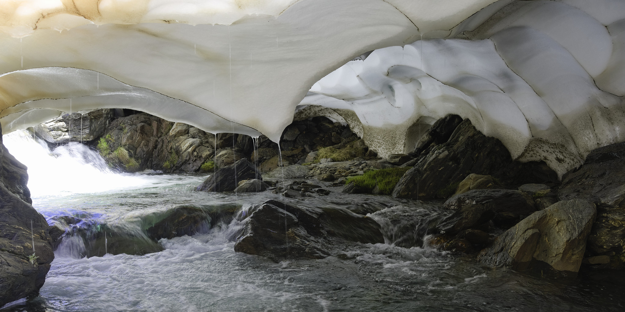 A river runs below a huge cavern of snow. In the background is a patch of green shrubs emerging from their winter slumber. The roof is full of curving snow sculptures