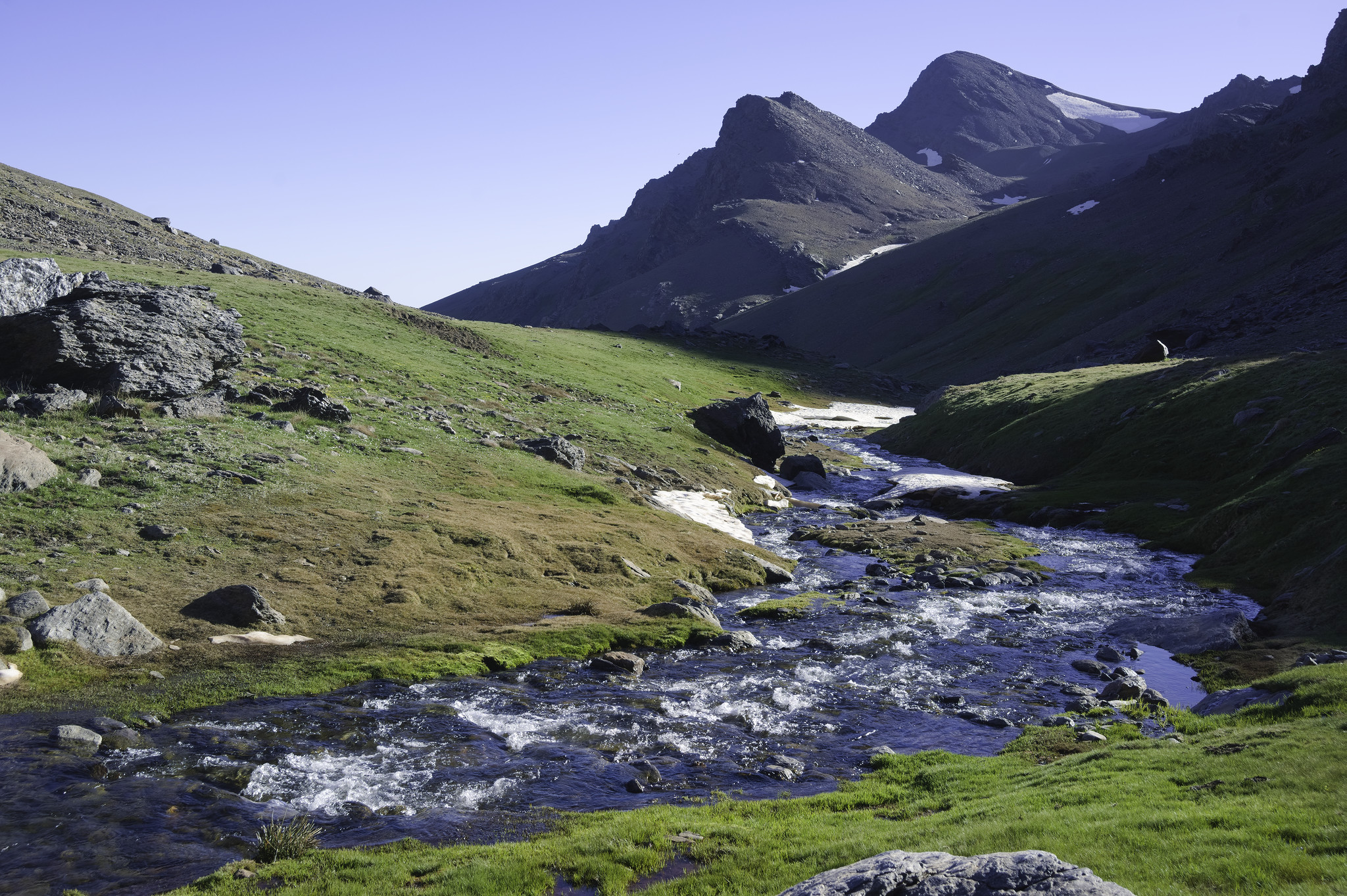 Green grasses follow a river as it curves round to the left on the right hand side of the image. The right hand banks of the river are in evening shadow. Ahead and to the right are some high mountains and some snow can be seen.