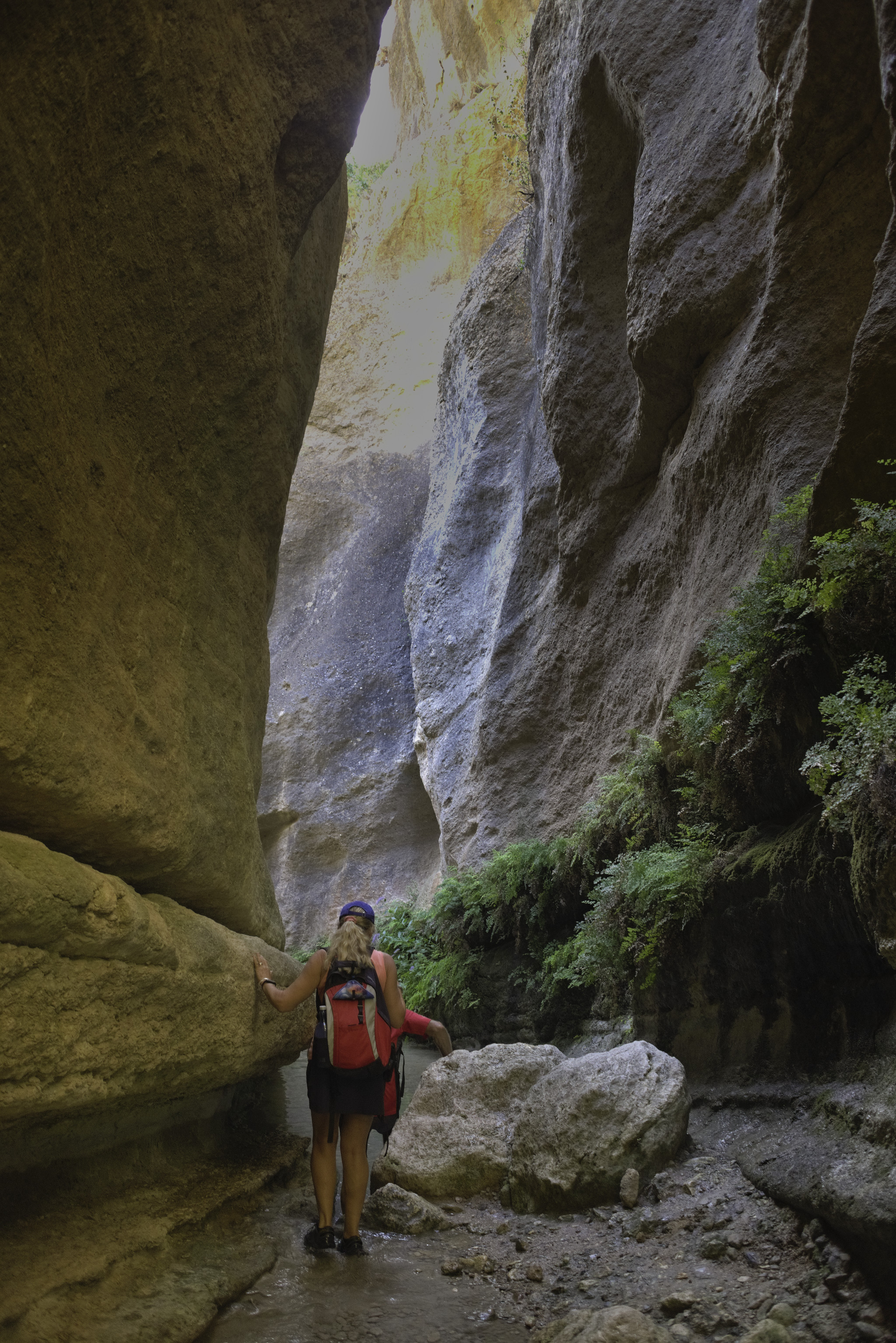 A hiker walks alongside in a shallow stream whilst huge rock colorful walls tower above