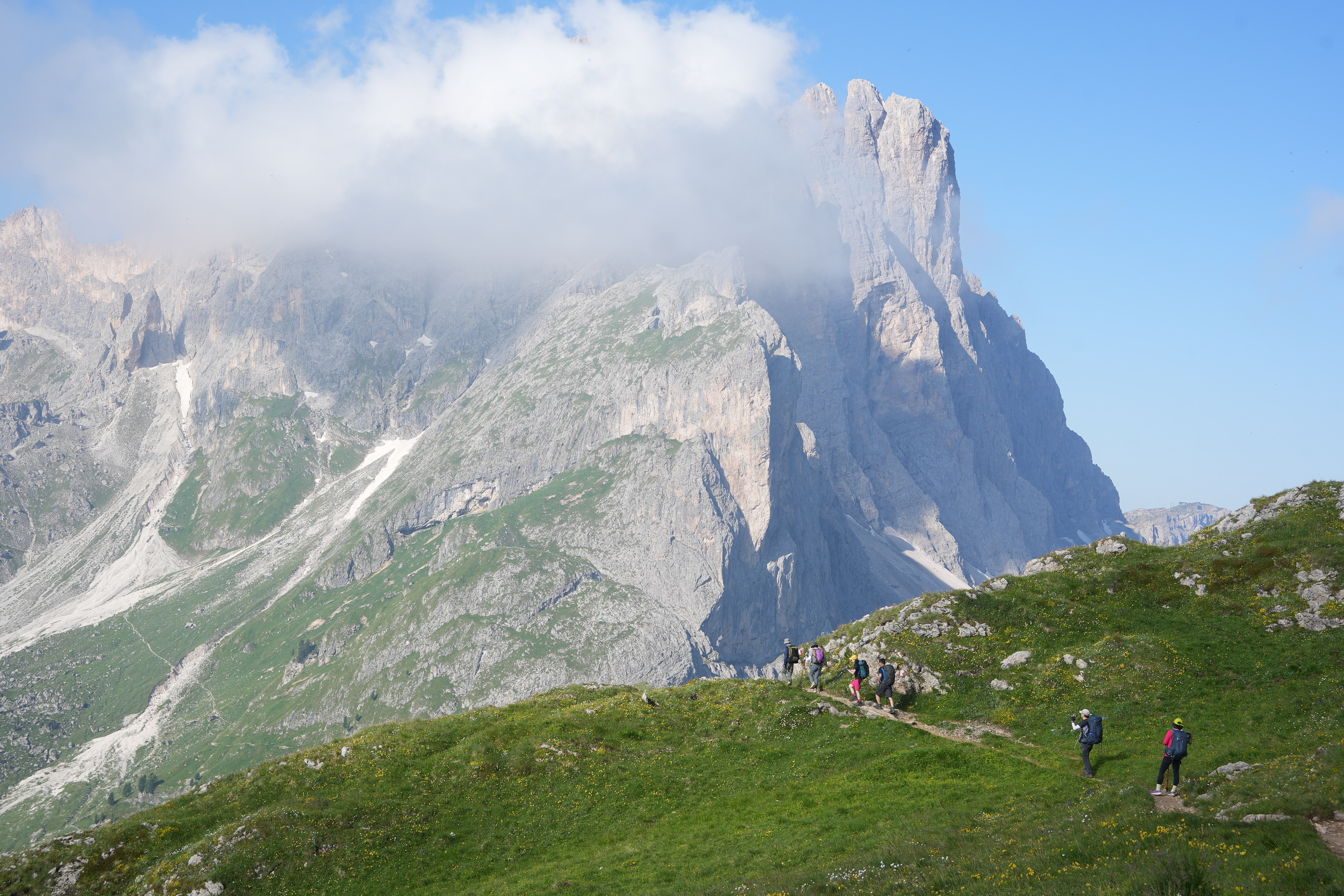 A mountain in cloud towers above some green meadows with some walkers