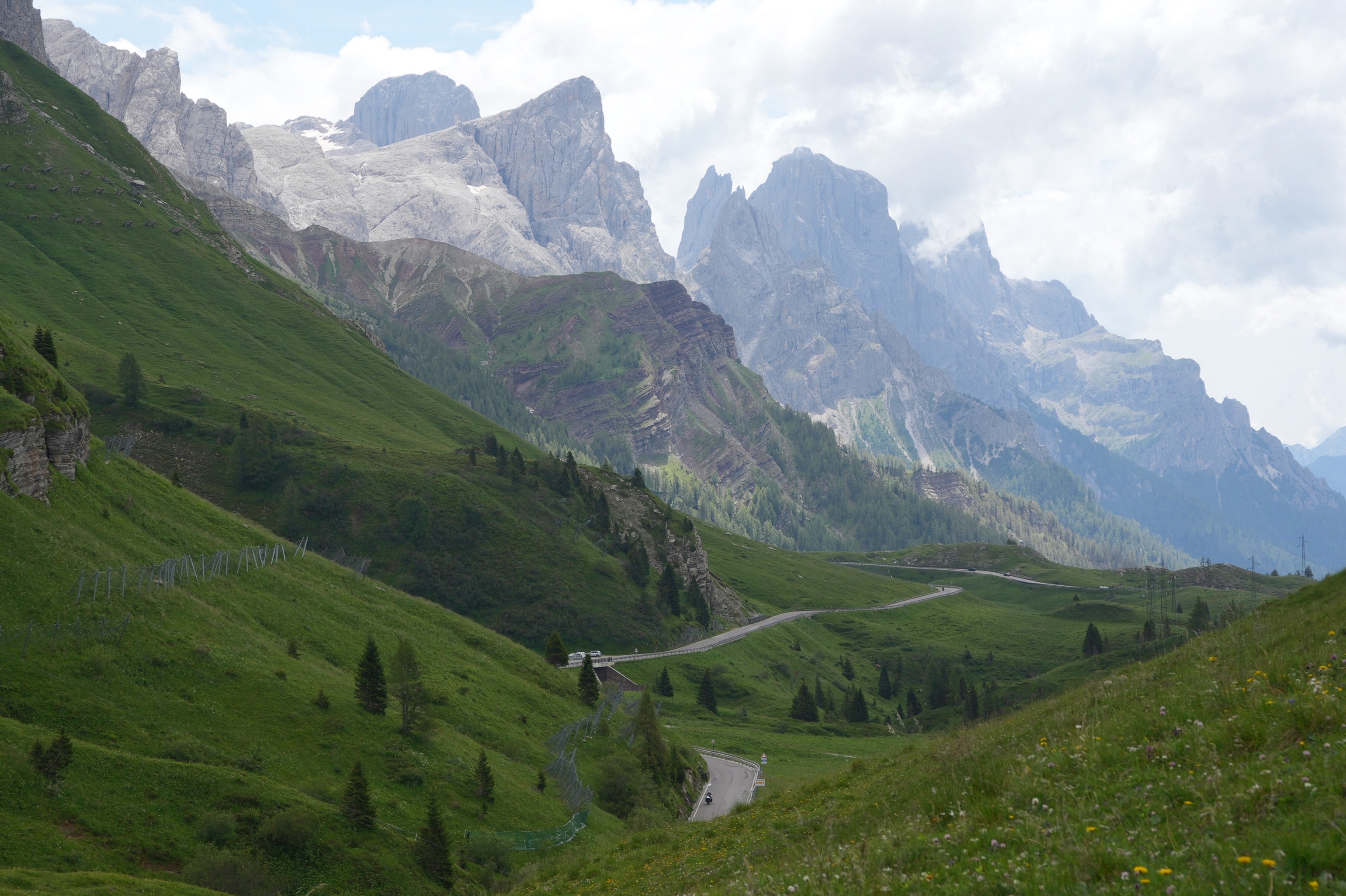 A road winds its way through green hillsides. Above rise some dramatic mountain peaks of the Pale di San Martino range