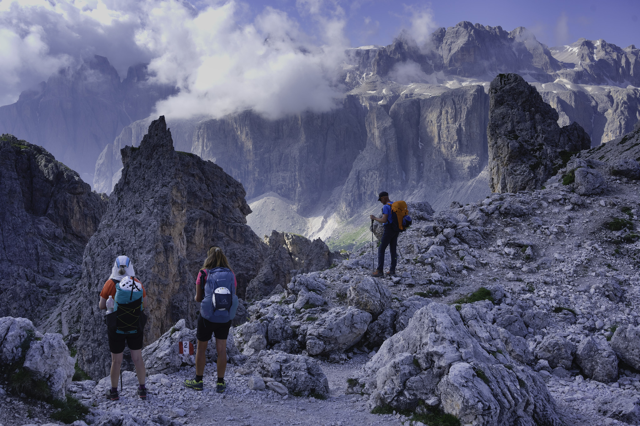 Three people gaze out at some sharp mountain peaks and pinnacles. In the background is another group of higher mountains with some cloud gathering around the summits