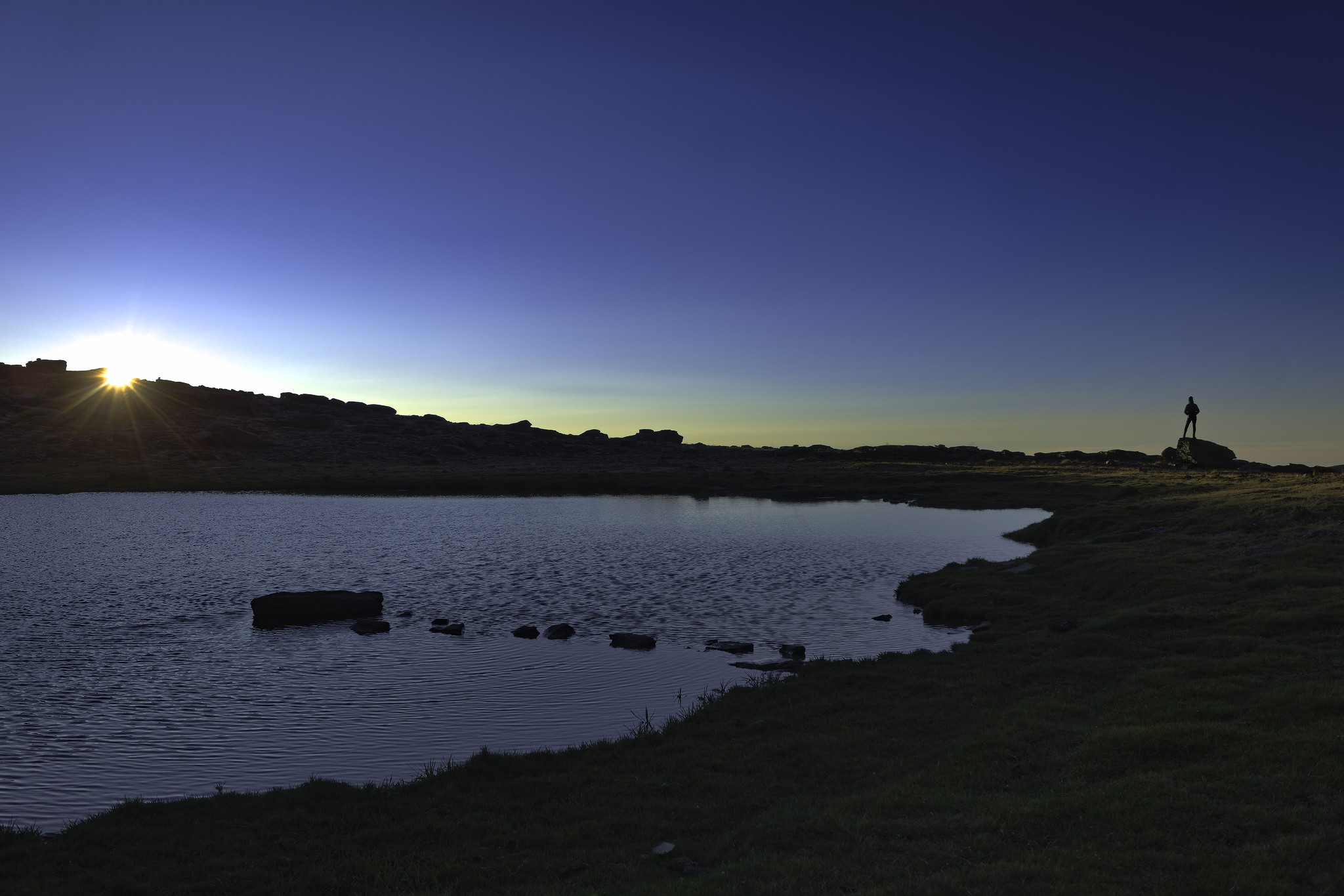 The morning sun appears over a ridge line to the left whilst a person stands on an isolated boulder on the right. Nearer the camera there is a lake rippled with wind driven wavelets. The lake had some small stepping stones