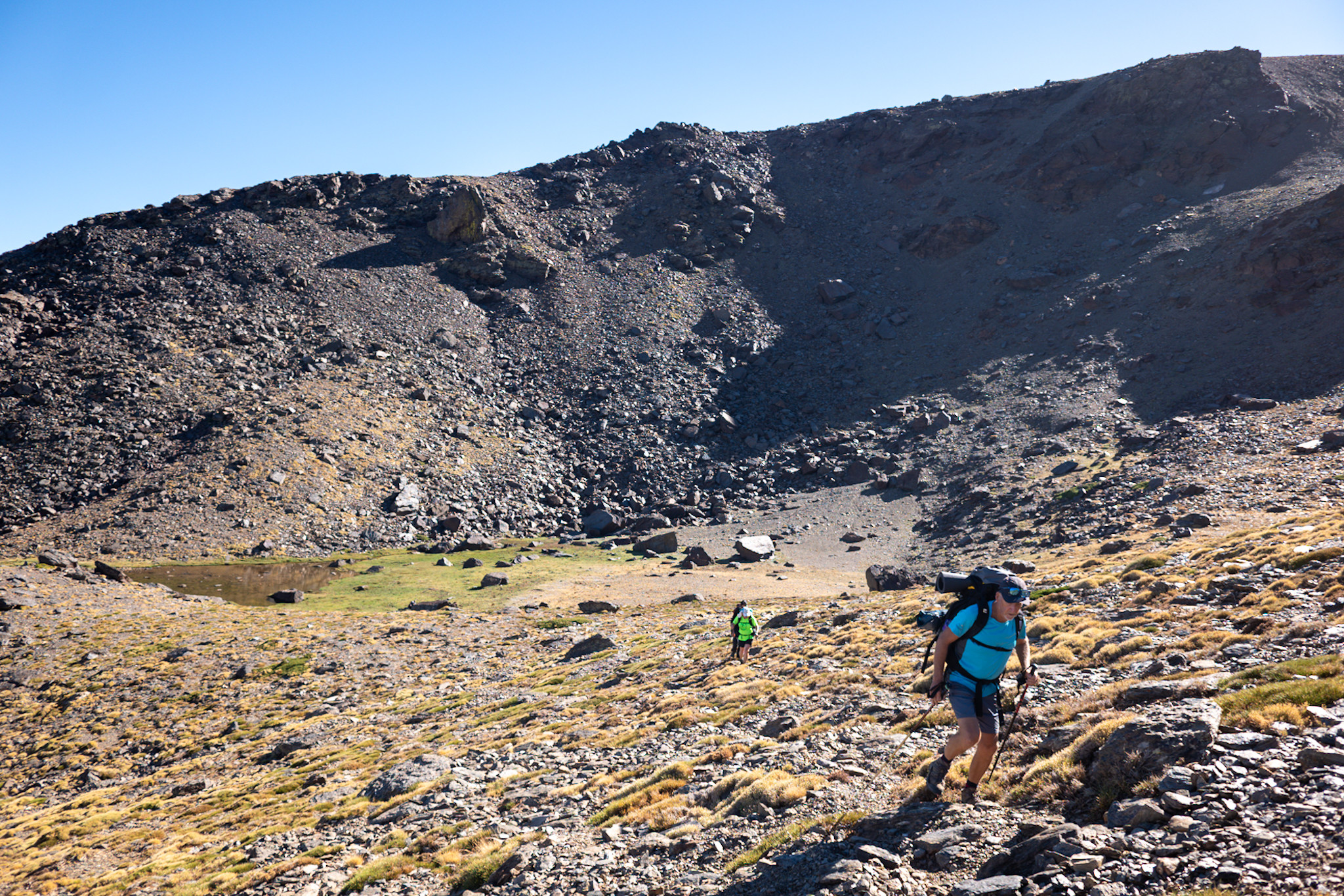 Two hikers leave a small lake. Big mountain walls behind