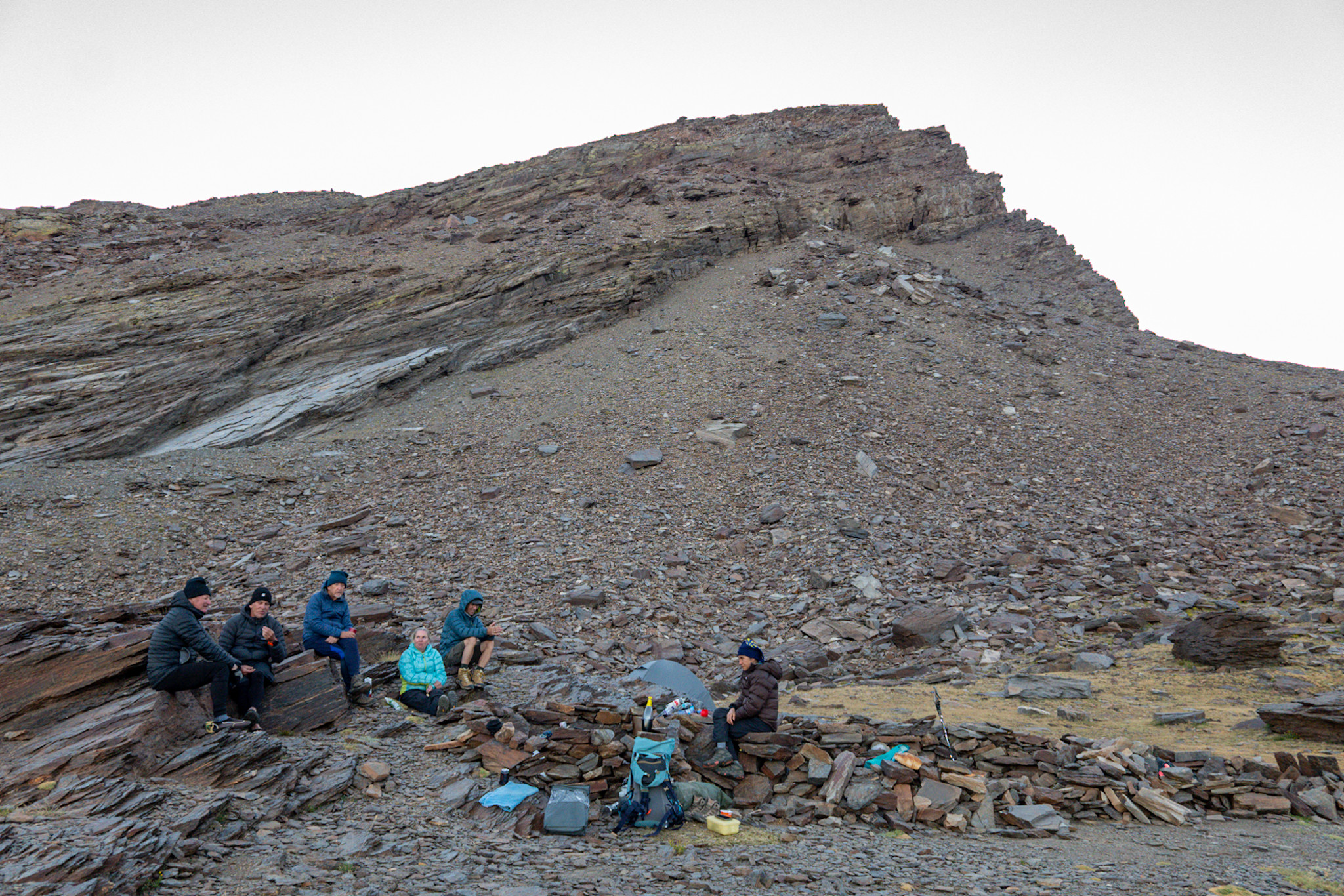 A group of backpackers bivouac in small walled shelters below a rocky mountain