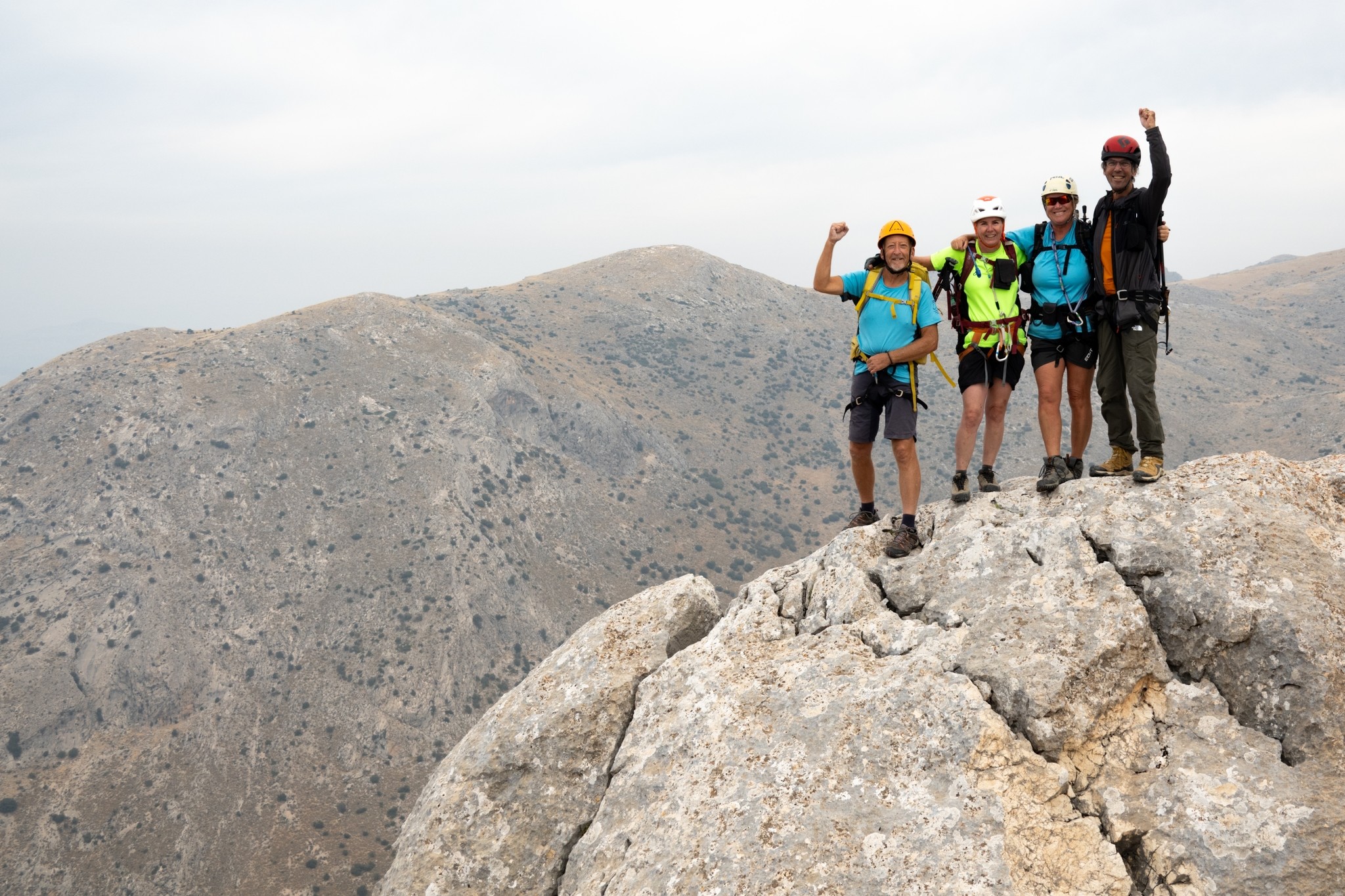 4 people with helmets on stand at the top of a grey rocky mountain