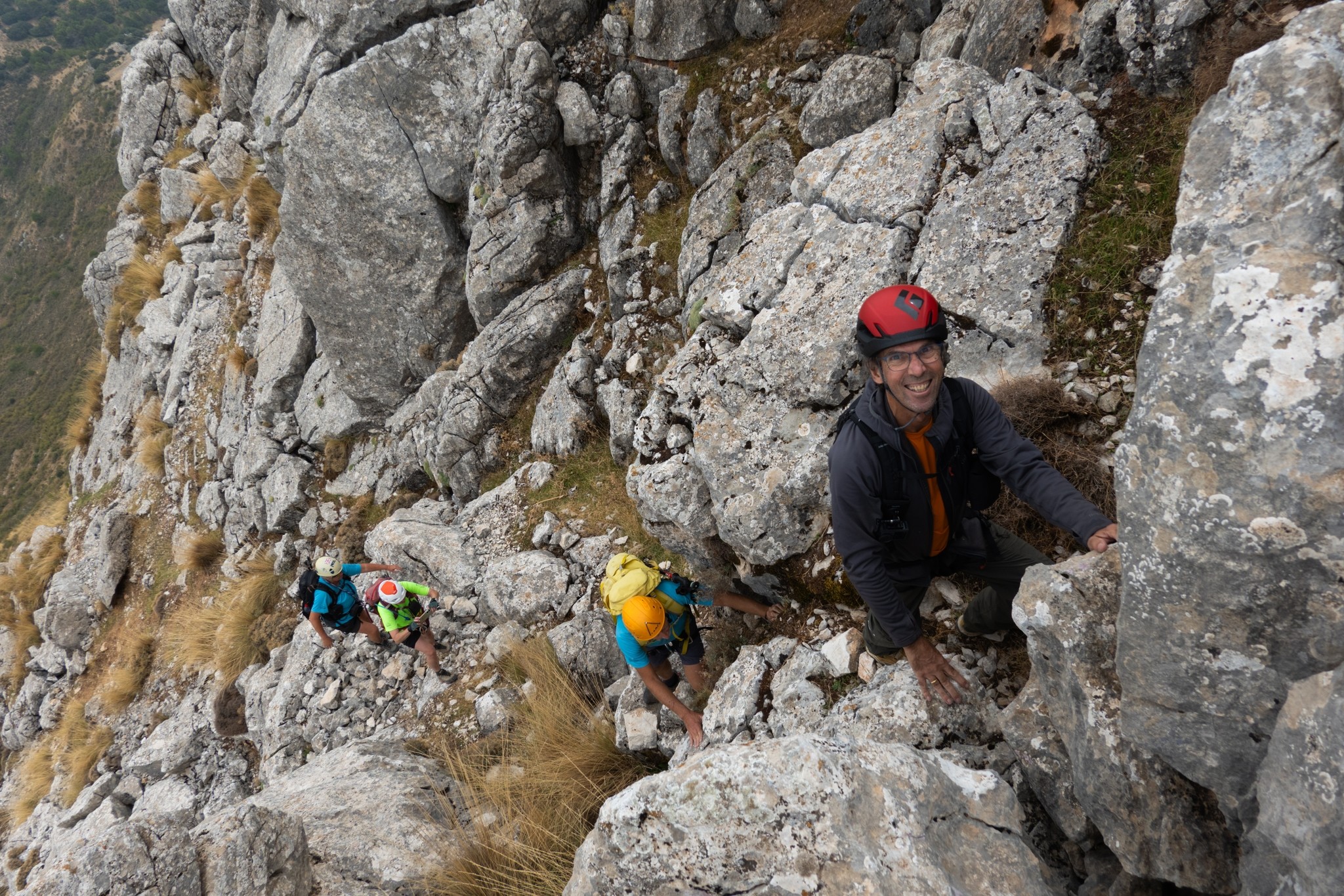 A person with red helmet looks up at the camera whilst others make their way up to him. They are climbing a rocky hillside