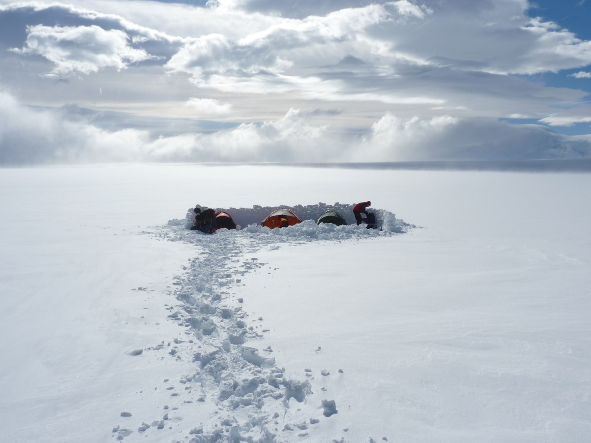 Aa small snow built shelter sits amid a white, flat landscape. The shelter contains 3 small tents