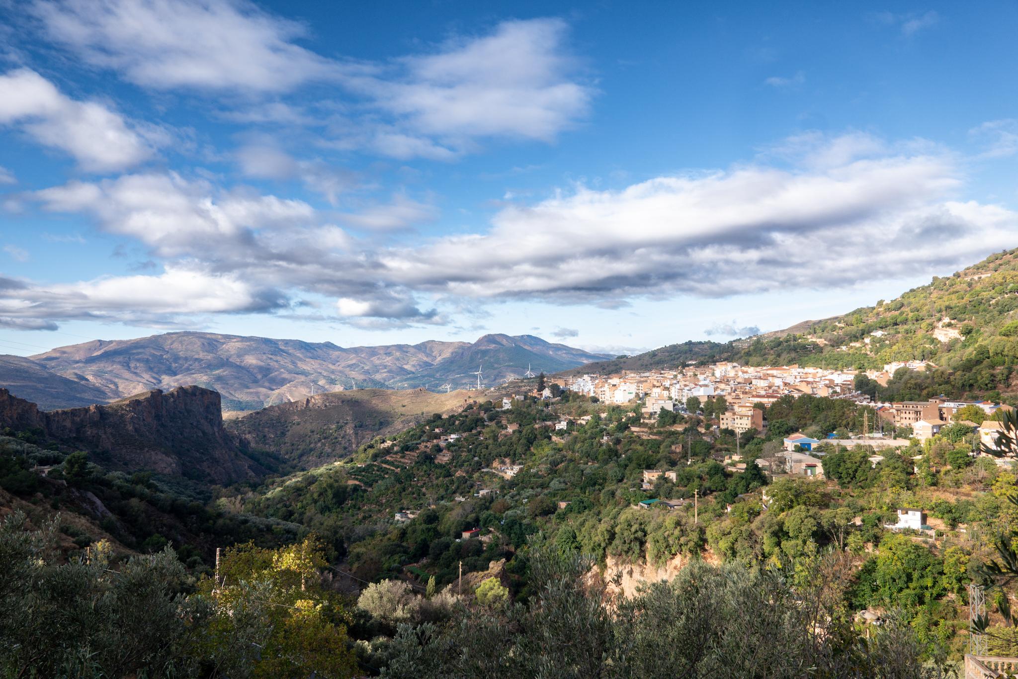 A small town sits on a small plateau above a valley. Above the town are blue skies and whispy clouds