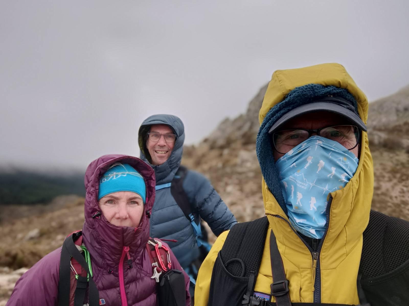 Three cold looking hikers are wrapped up against the cold biting wind in the mountains