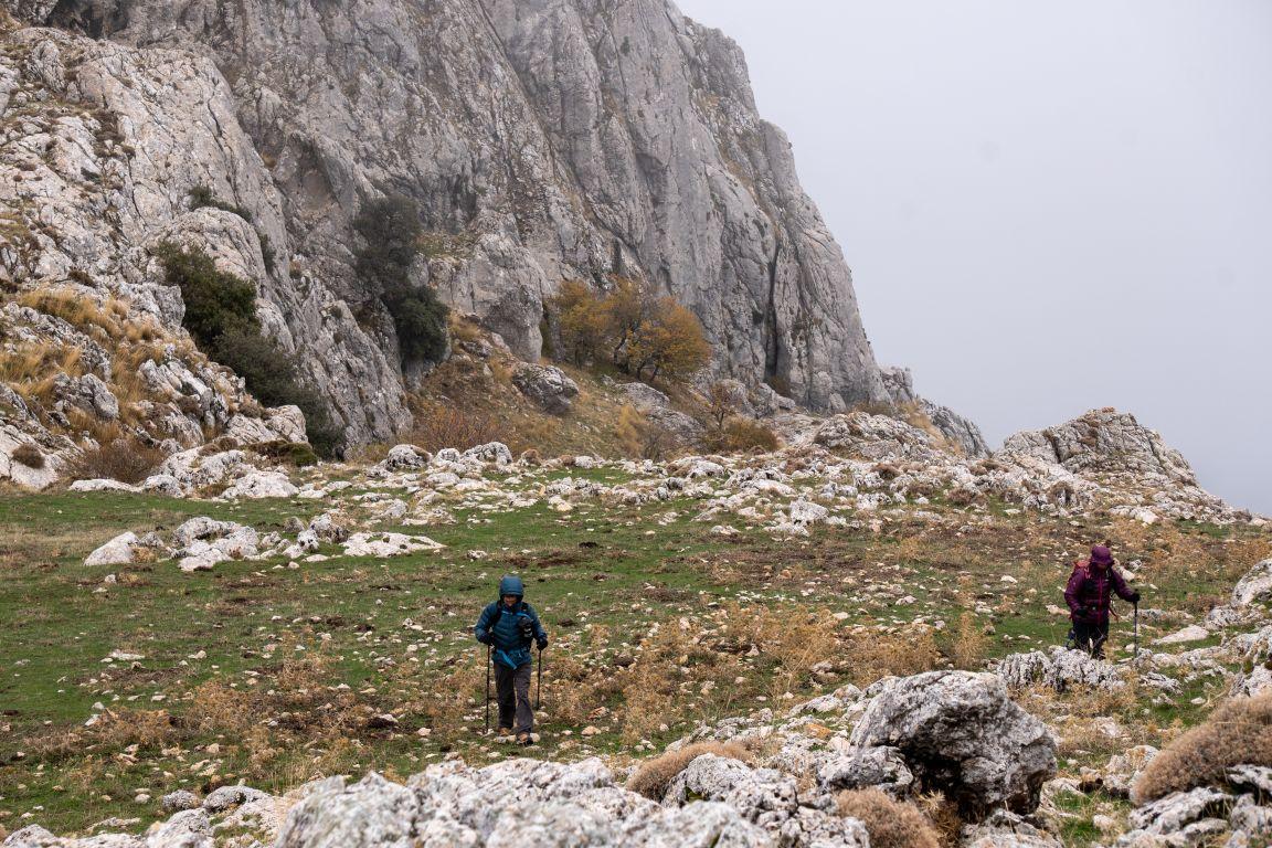 Two hikers cross a stonry pass with a big crag behind them