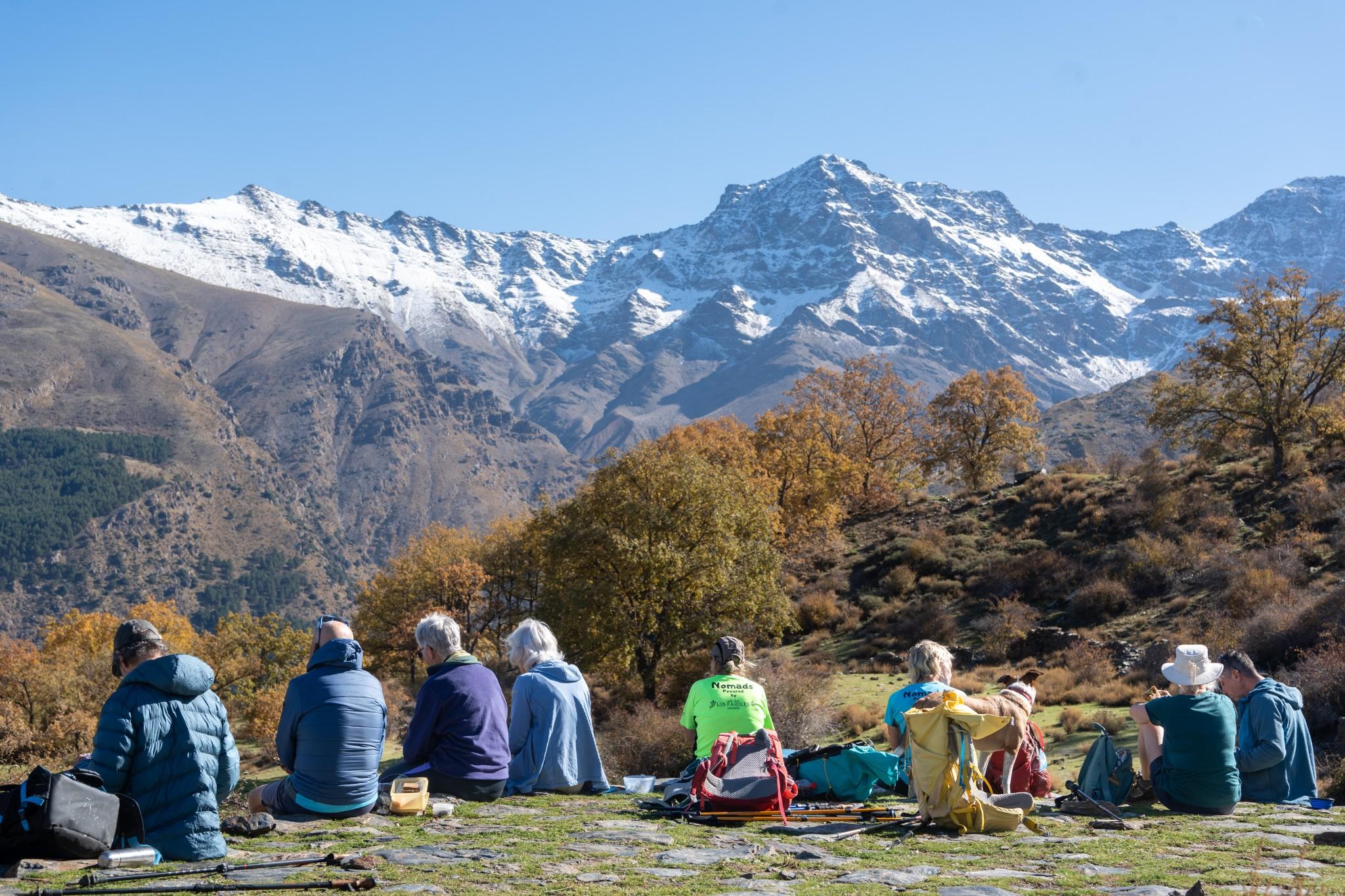 A group of hikers sit on a flat grassy area looking at a view of snow capped mountains