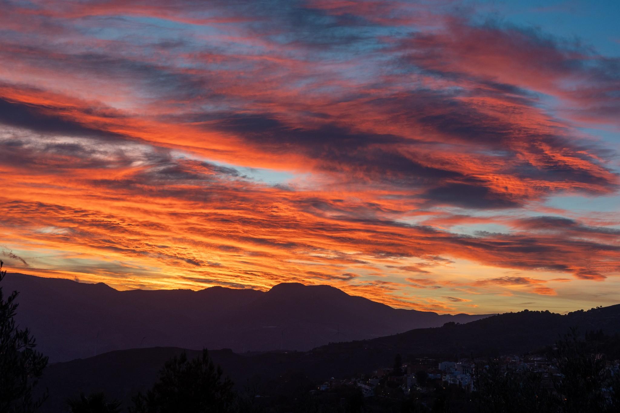 A multi coloured sunset with patches of light blue sky behind. A faint outline of a town in the lower third of the image.