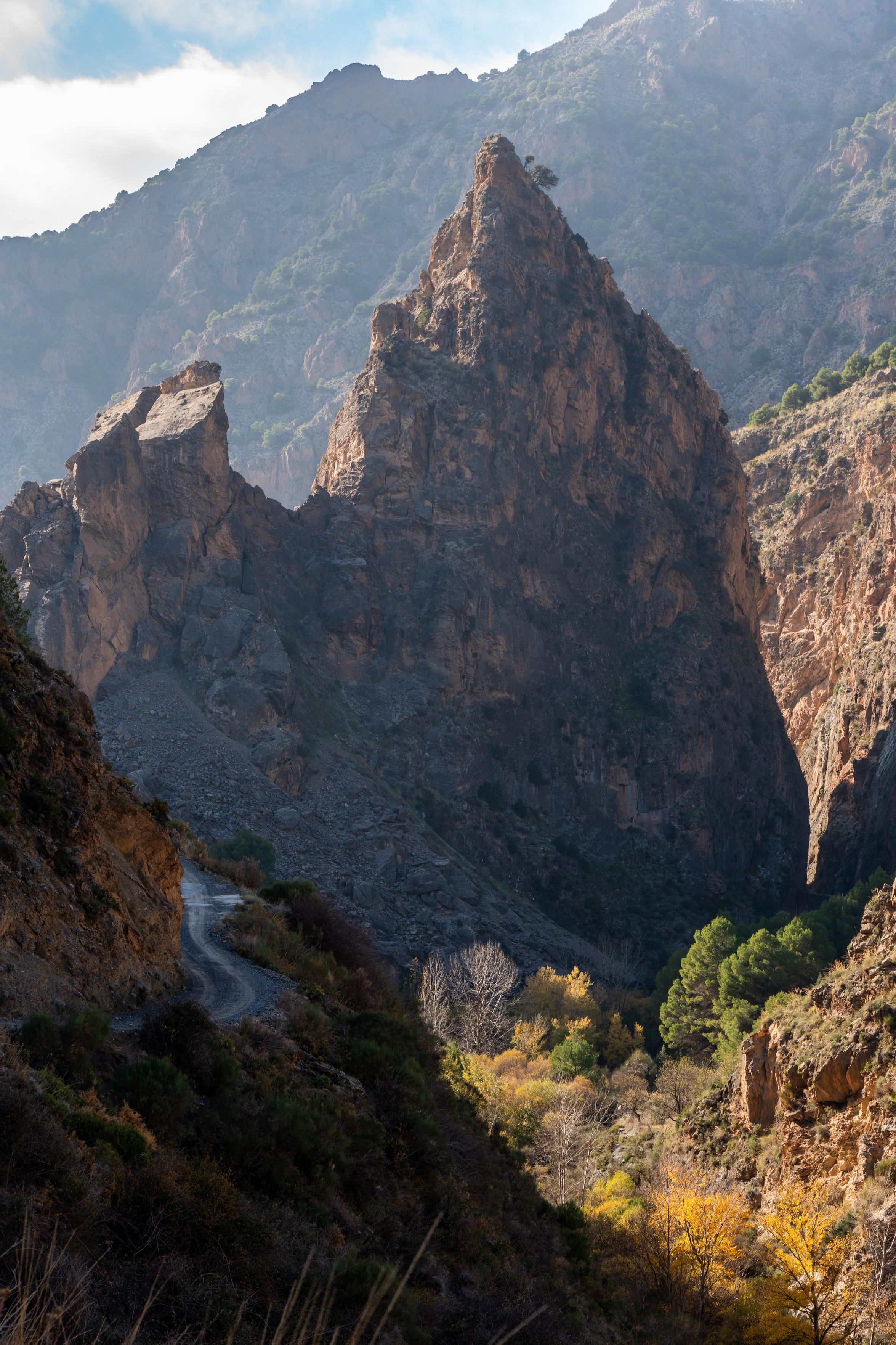 A dramatic gorge illuminated with autumn colors. To the left runs a small road alongside the hillside. A sharo peak rises in the center