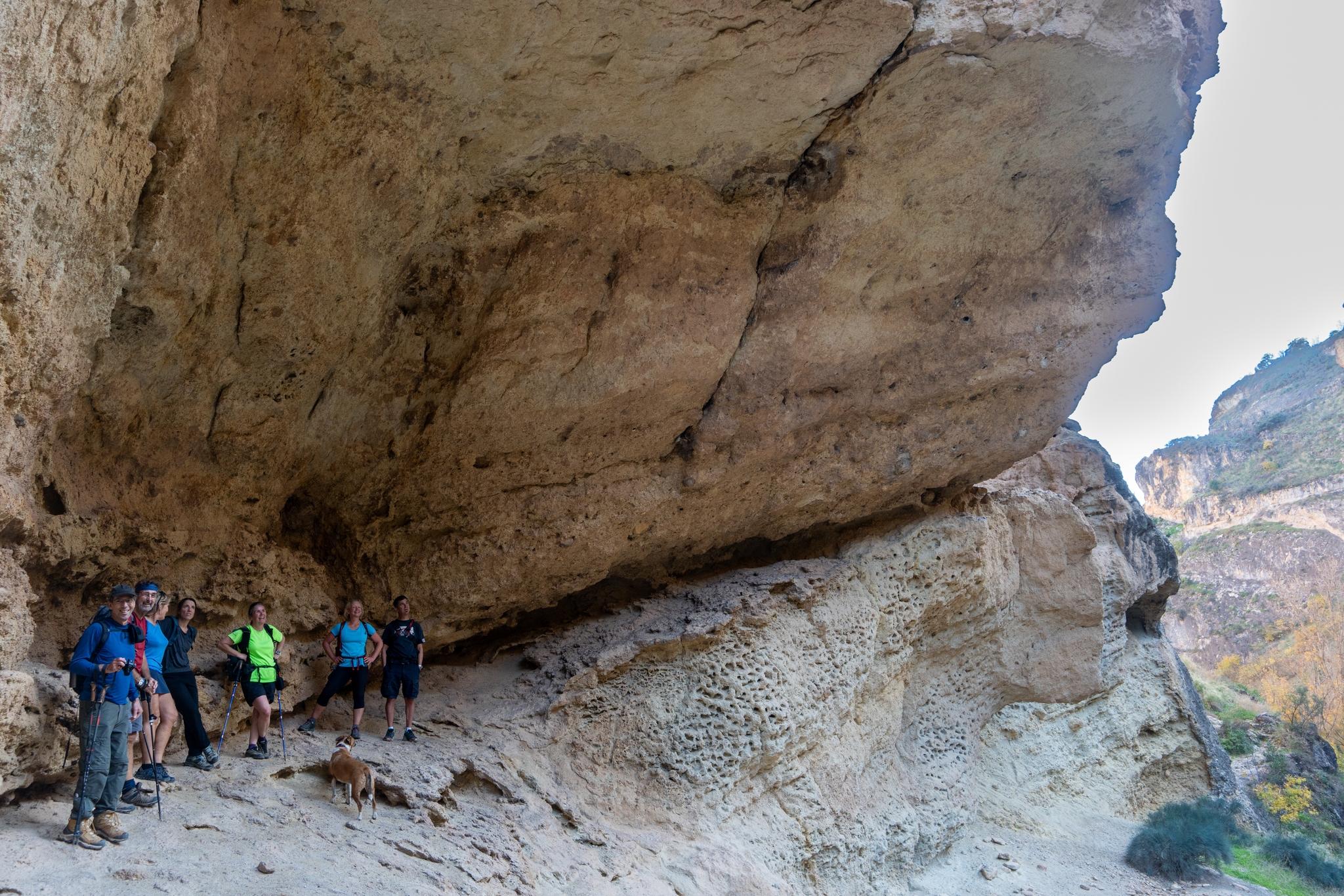 a group of hikers shelter in the shade beneath a huge overhanging cliff