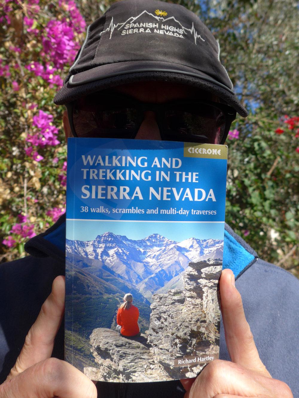 A person with a baseball cap on and dark glasses has a mountain guidebook held up in front of his face