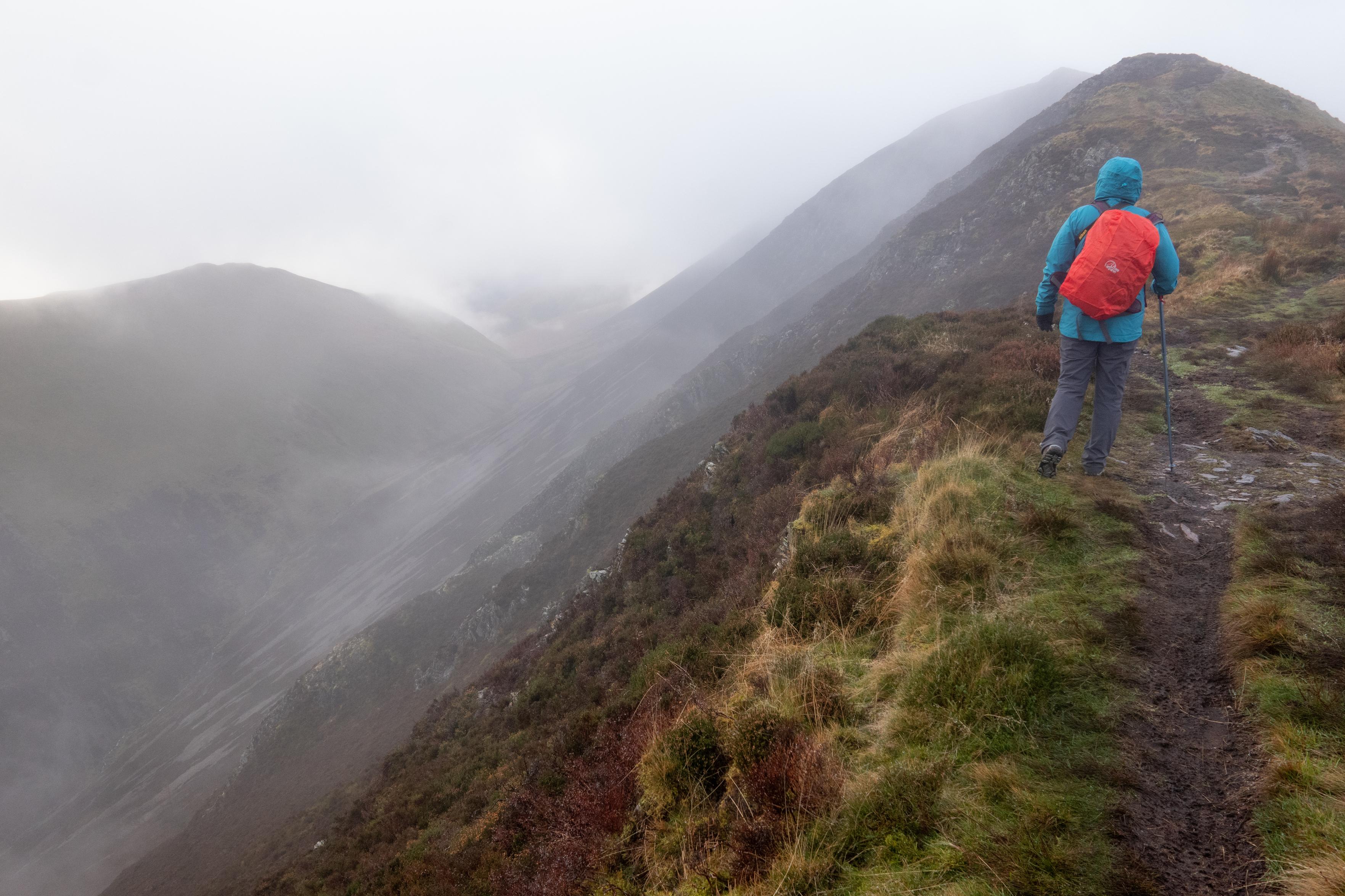 A person in blue jacket and red rucksack ascend a broad ridgeon the right. To the left lie mountains mists and ridges