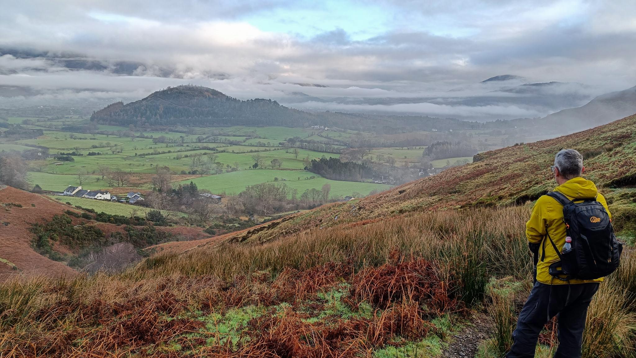 A person in yellow jacket on the right looks down Coledale Beck towards the Newlands Valley and Keswick, Derwentwater area. Green fields mix with bright orange slopes of heather and braken.