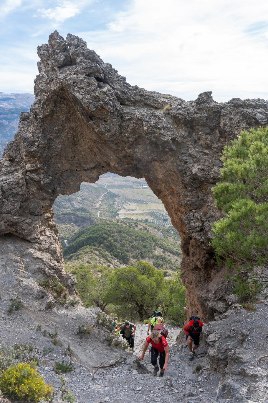 A rock arch forms a window to lower ground and olive groves. Some people climb up through the window