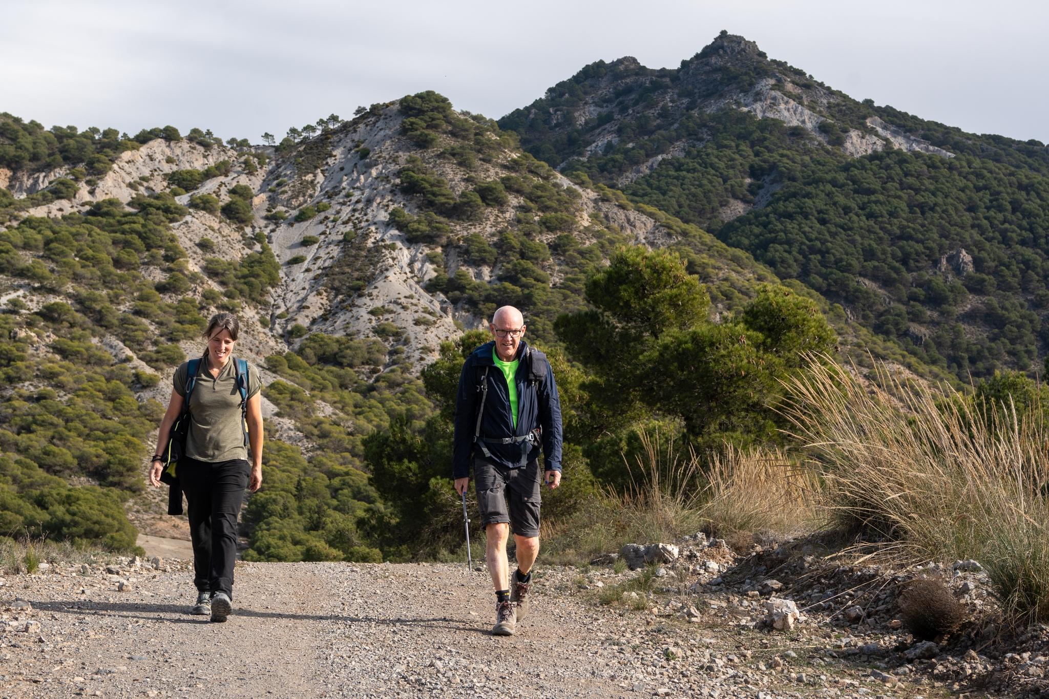 Two hikers walk down a dirt track and away from the tree covered peak of Silleta de Padul