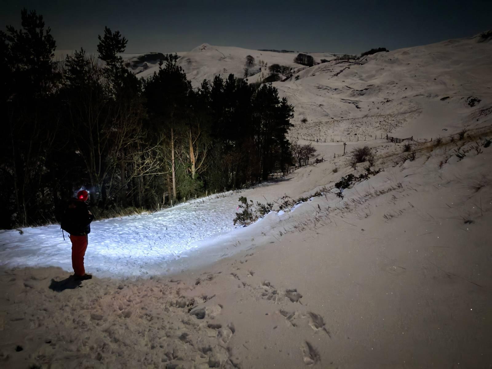 A person stands at the side of a dark forest at night. The way ahead is illuminated by his headtorch. The snows lead up to a broad mountain peak