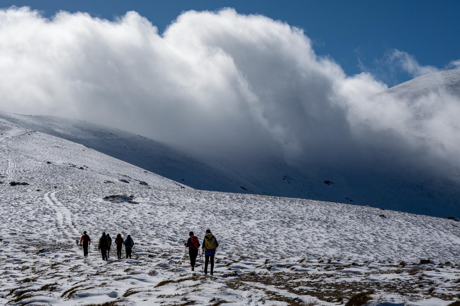 Hikers walk along a snowy track to the left. On the right a huge bank of cloud appears rolling over the mountains