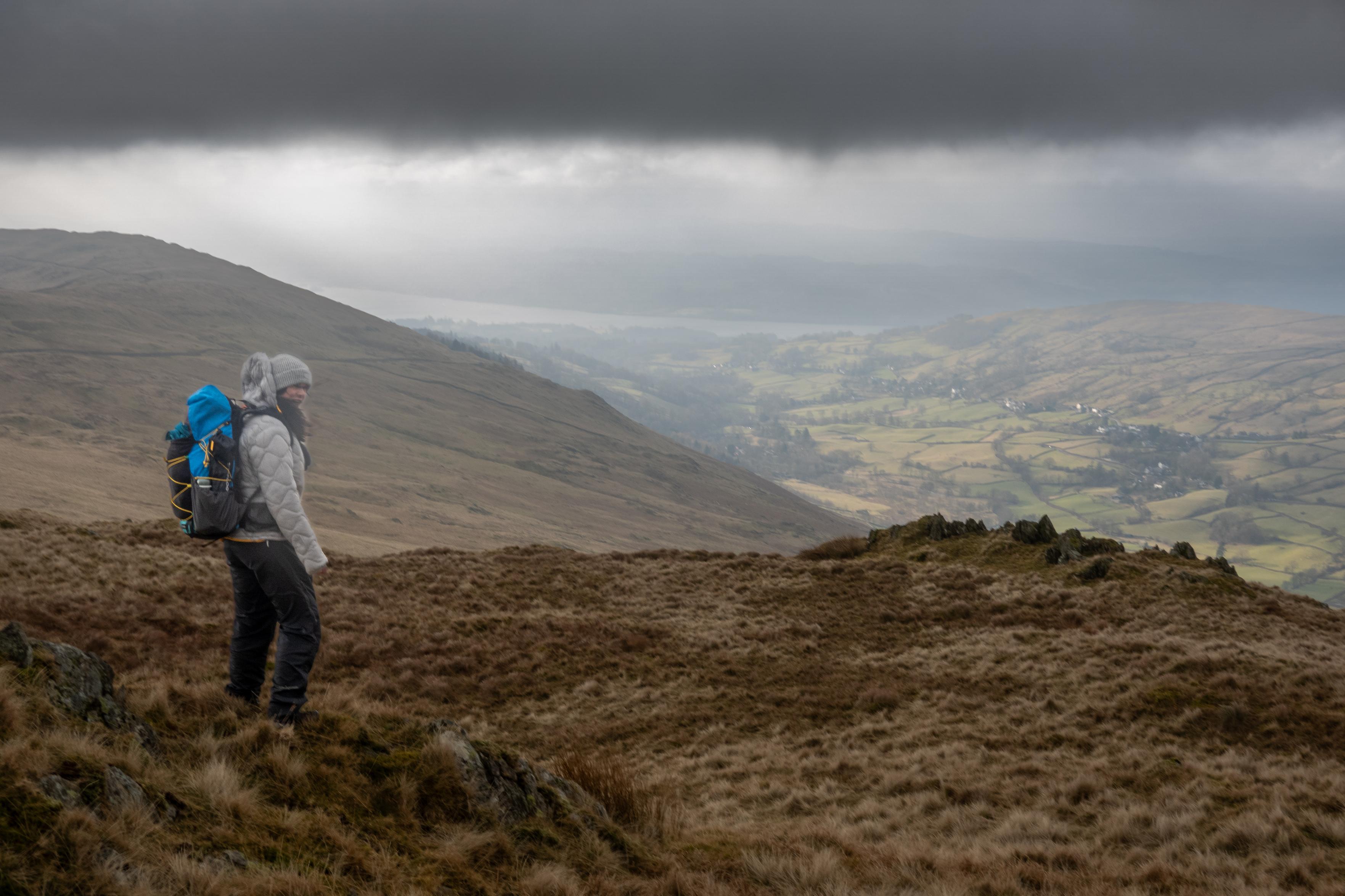 A person stands on a rocky outcrop. It's a dull day with dark clouds above but in the distance there is the intimation of sun