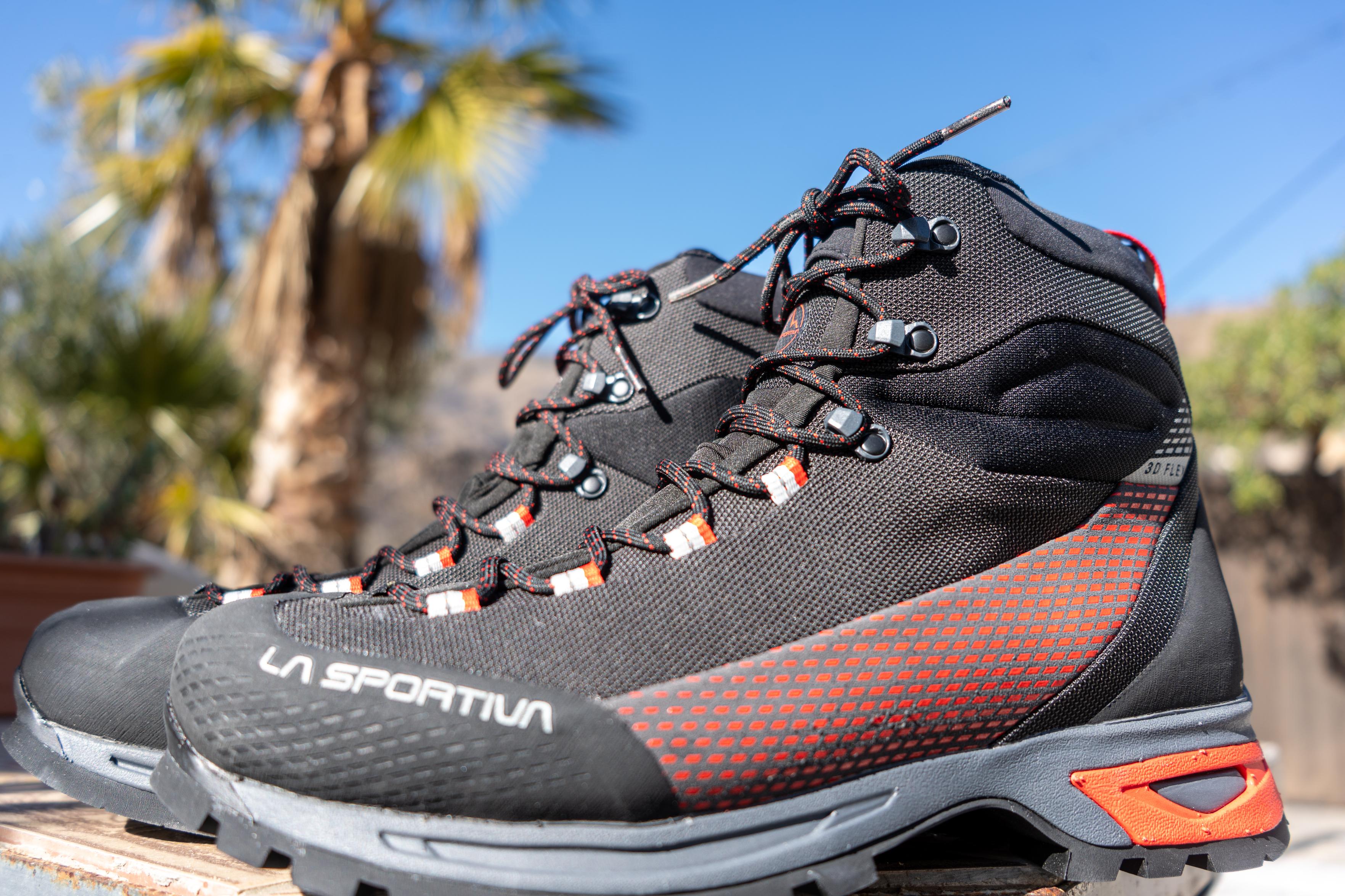 A pair of hiking boots from La Sportiva  stand side by side on a table. Above is blue sky and a palm tree
