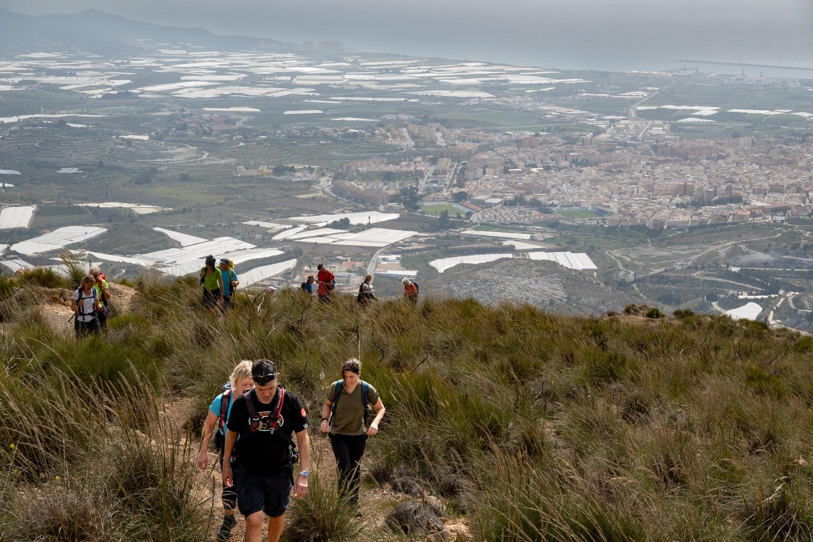 Nearing the summit, a group of hikers pass their way round low shrub land. behind and far below is the city of Motril, Spain