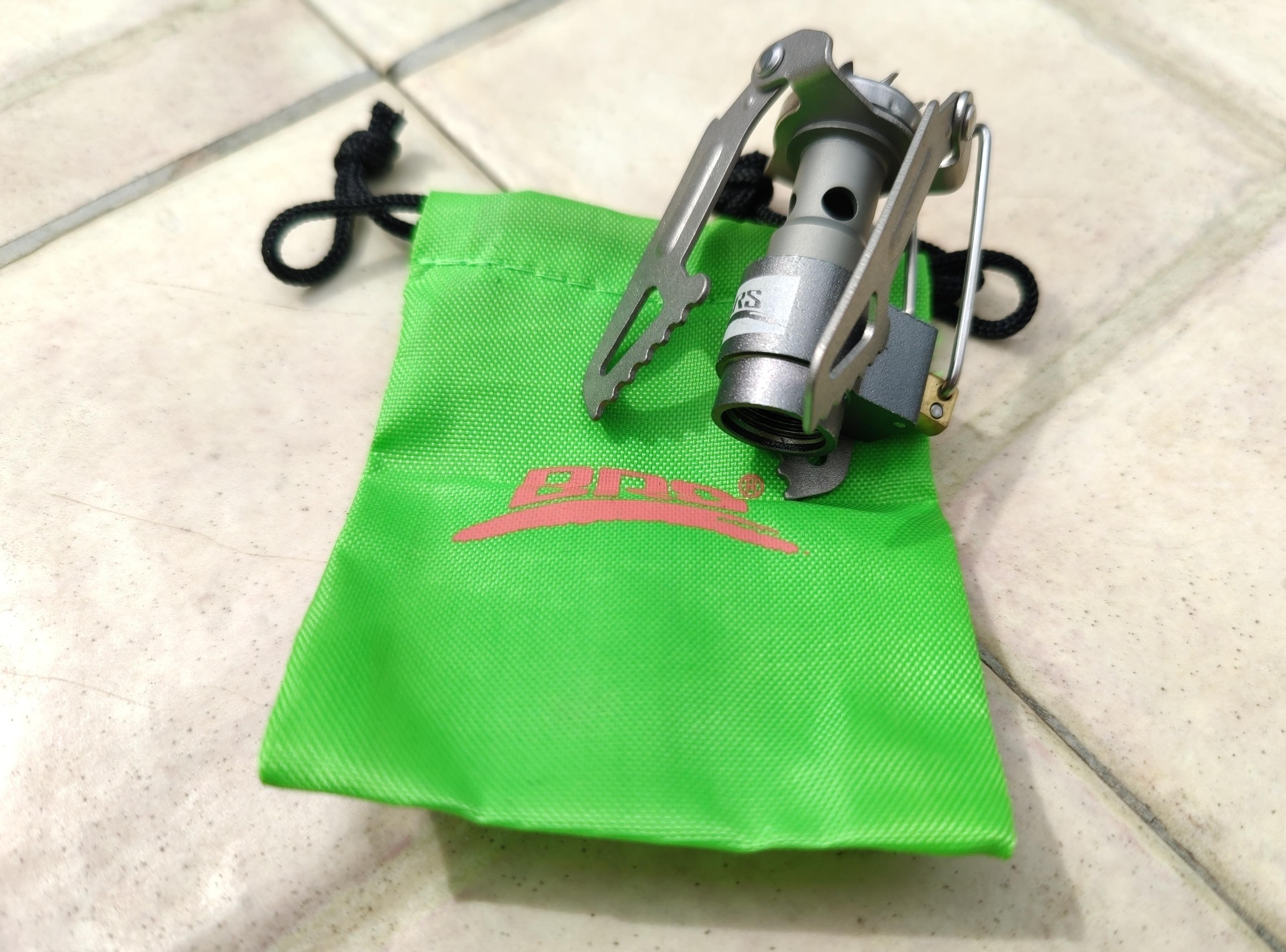 A titanium camping stove sits on a green bag