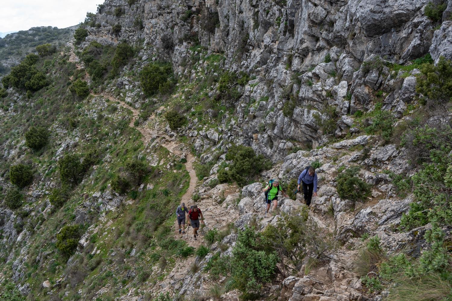 A narrow mountain track which is starting to rise amongst rocky outcrops. Some walkers are on the track and a huge grey wall of rock lies to the right