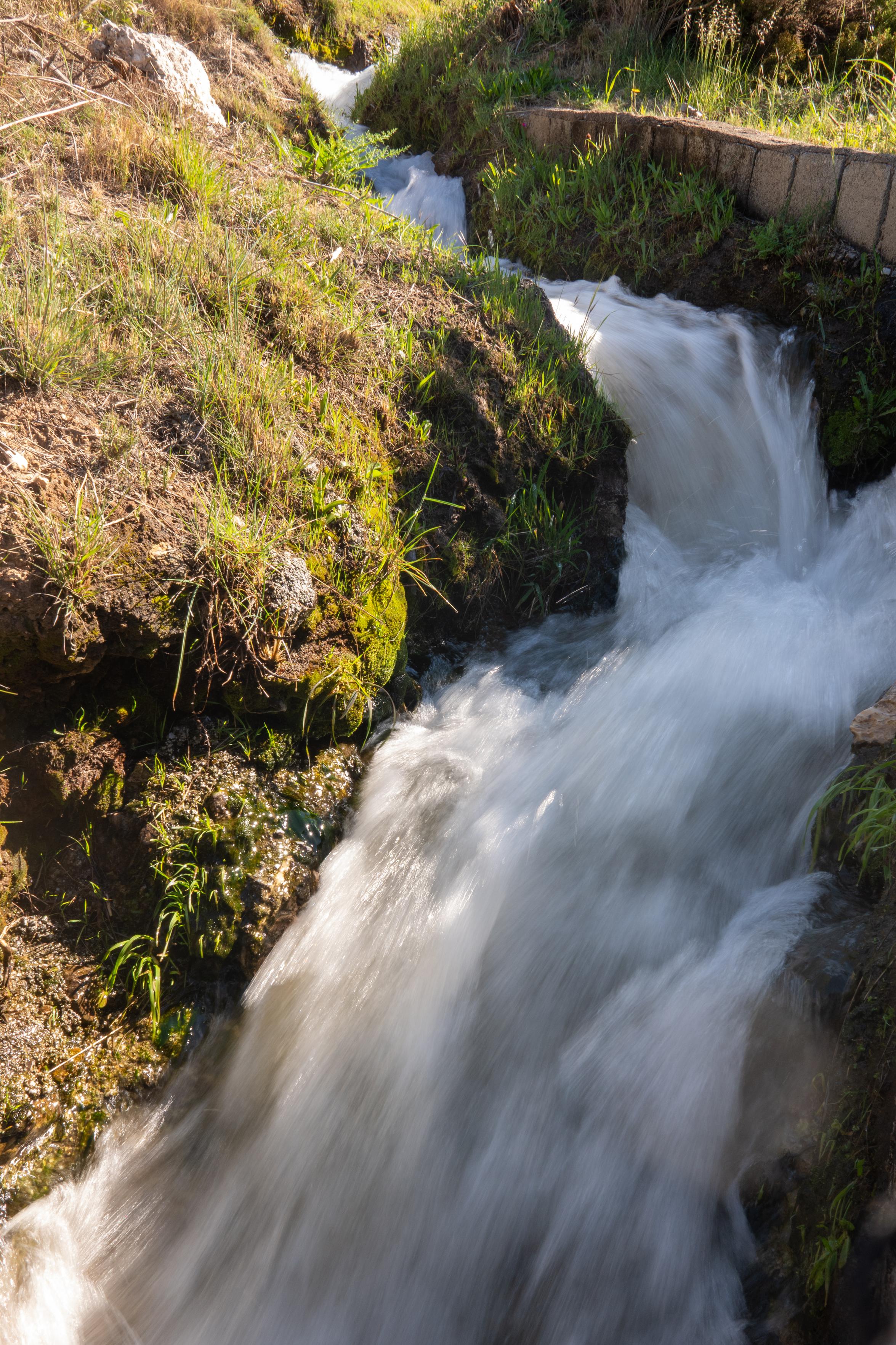 Waters plunge down an irrigation channel with green grassy banks and a short wall top right
