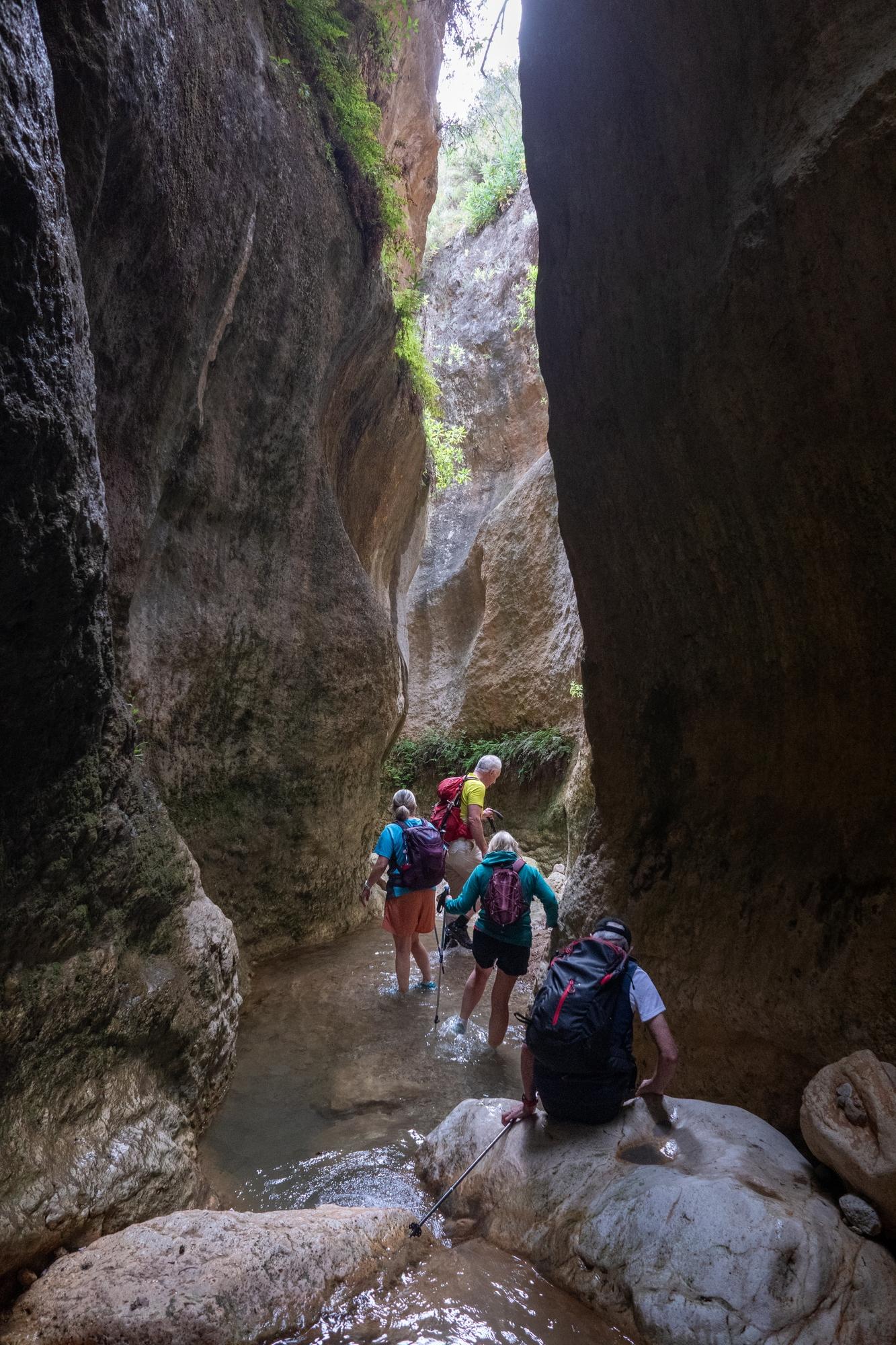 A group of hikers passes down a narrow river gorge with huge rock walls vertically rising above
