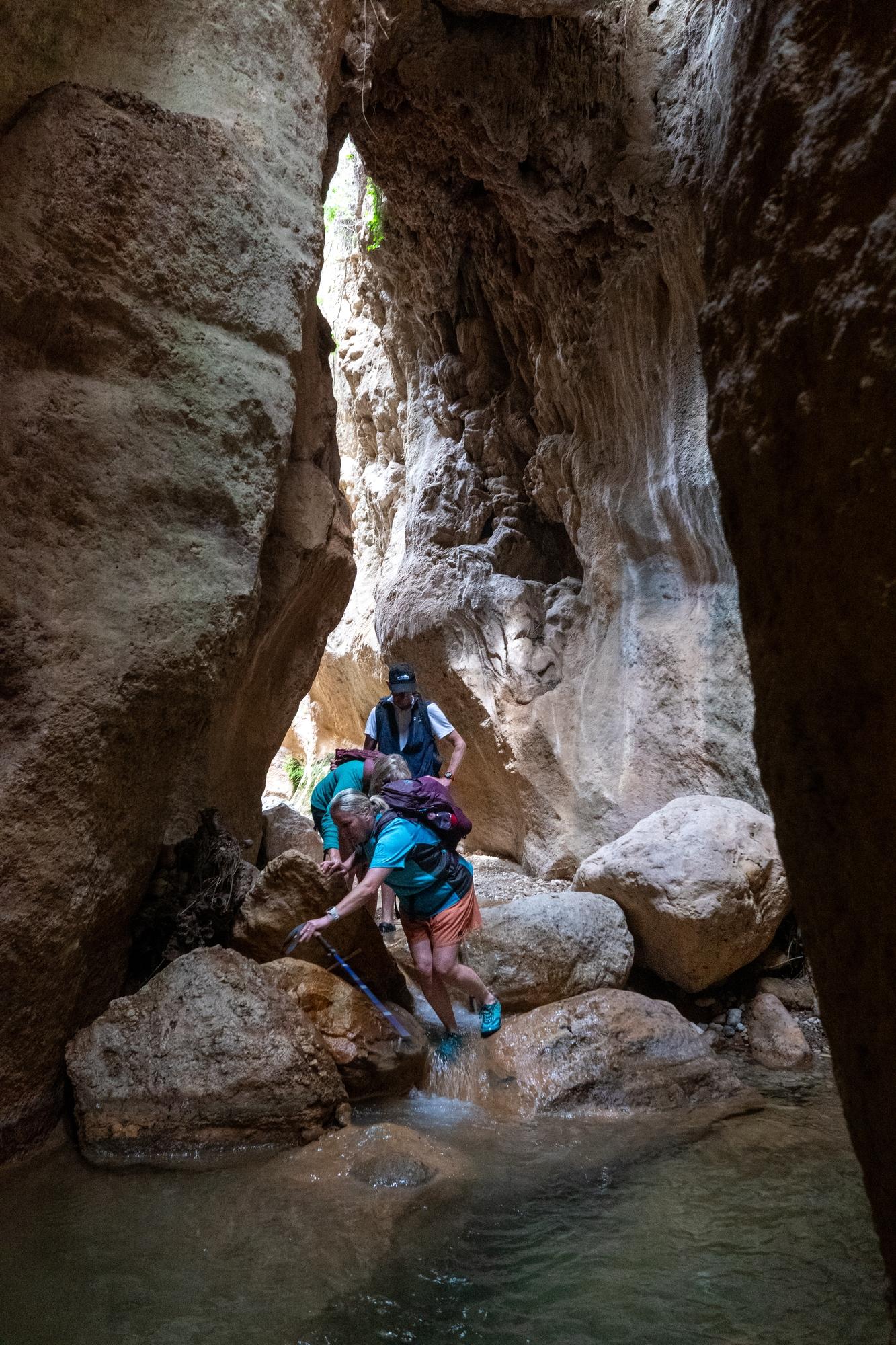 A group of hikers passes down a narrow river gorge with huge rock walls vertically rising above