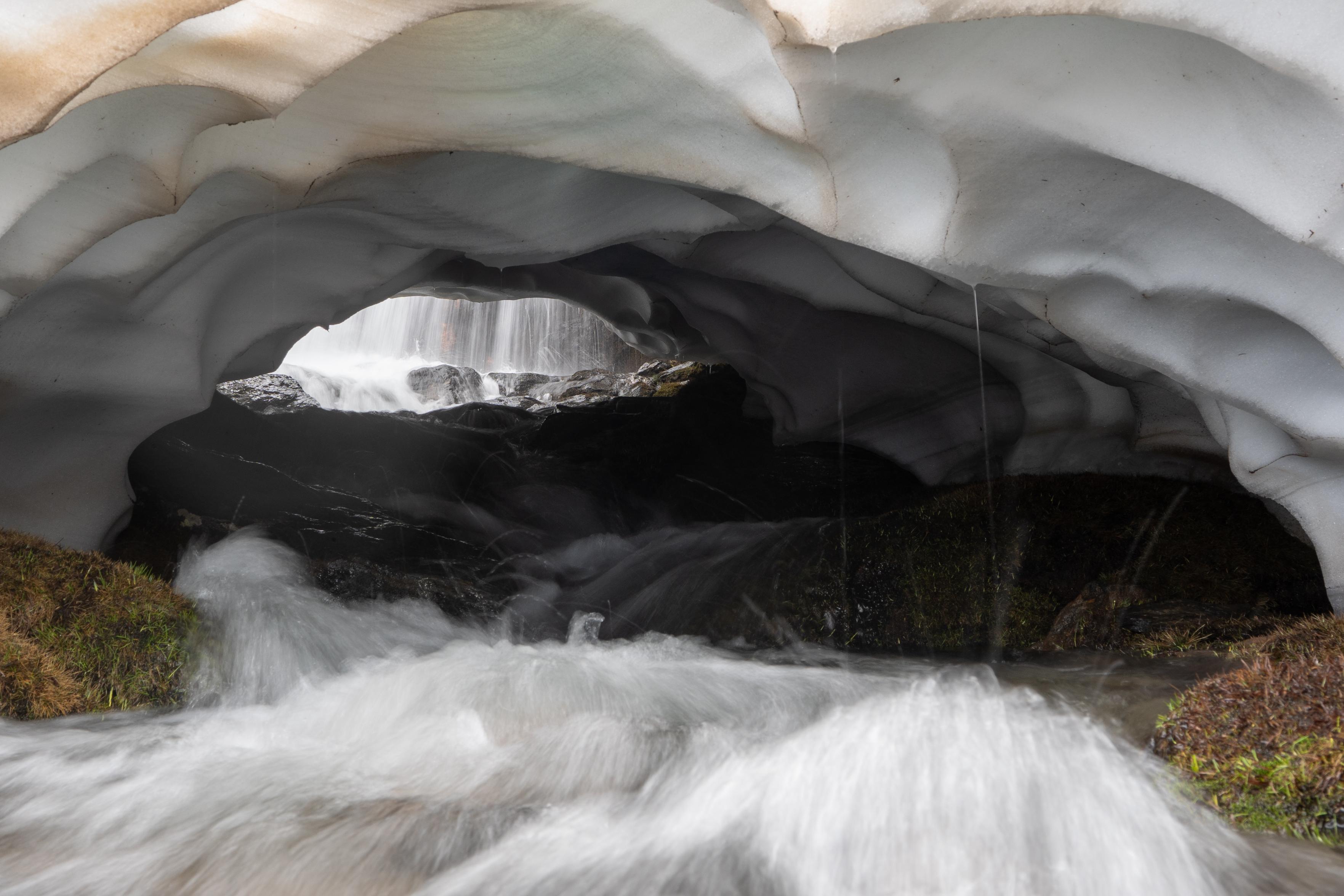 A tunnel underneath the snow where a fast flowing river flows. There is light at the far end of the tunnel where a waterfall can be seen.