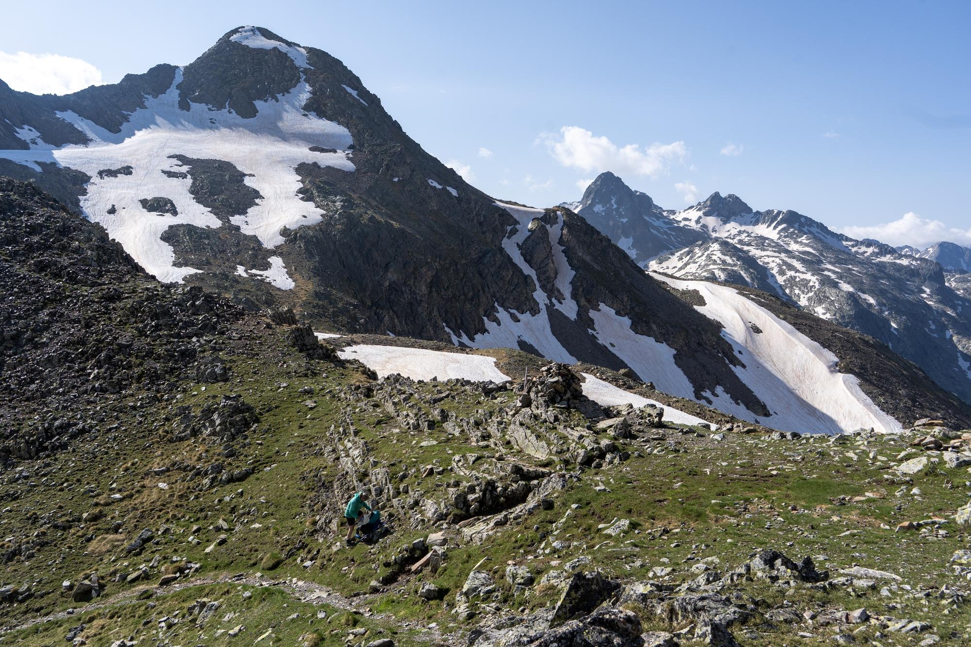 A green and level mountain pass has snow clad mountains rising behind. A small figure with rucksack in lime green can be seen near the pass